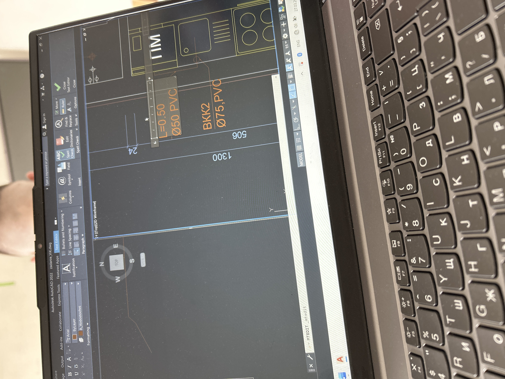 A close-up view depicts a grey laptop, angled downwards, displaying Autodesk AutoCAD 2022 software on its screen. The title bar indicates an active file named "zadanie_VIK.dwg" and the "Text Editor" tab is selected. The CAD drawing shows architectural or engineering plans with dimensions such as "24", "1300", and "506". Specific elements include pipe specifications: "L=0.50 Ø50 PVC" and "BKK2 Ø75,PVC," suggesting a water supply and sewerage (VIK) system diagram. A navigation cube showing "TOP," "N," "W," "S," and "E" is visible. The command line displays "MTEDIT _mtedit." The system tray shows "ENG" language and the date "27.03.20." Partially visible at the top edge of the screen is the blurred forehead and hairline of an individual, indicating an active user looking down at the display. The laptop's keyboard, visible in the foreground, features both Latin and Cyrillic characters, confirming a Bulgarian keyboard layout. The scene is located in Sofia, Bulgaria, likely within an office or home environment, suggested by the out-of-focus, light-colored background.