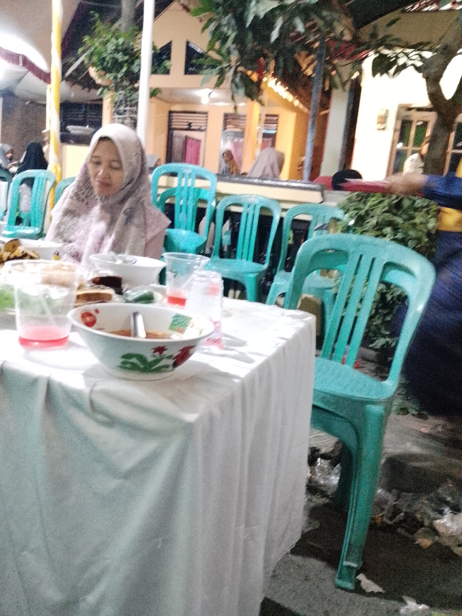 This is a medium-shot, outdoor photograph taken at night in Trangkil, Indonesia, likely during a gathering. In the foreground, a white tablecloth covers a table laden with food and drinks, including bowls of food, cups with red liquid, and possibly some cake. The food items are in white bowls with red and green floral patterns. In the midground, a woman wearing a light-colored hijab is seated, appearing to be looking down at the food. Behind her and to her sides are several bright turquoise plastic chairs, some occupied by unseen individuals. The background shows a building with yellow and brown accents, illuminated by warm lighting, suggesting an outdoor event or celebration. Trees with dark green foliage are visible, adding to the outdoor ambiance. The ground is dark and appears to be paved, with some scattered debris. The overall scene depicts a casual, communal dining event in a semi-rural or suburban Indonesian setting at night.