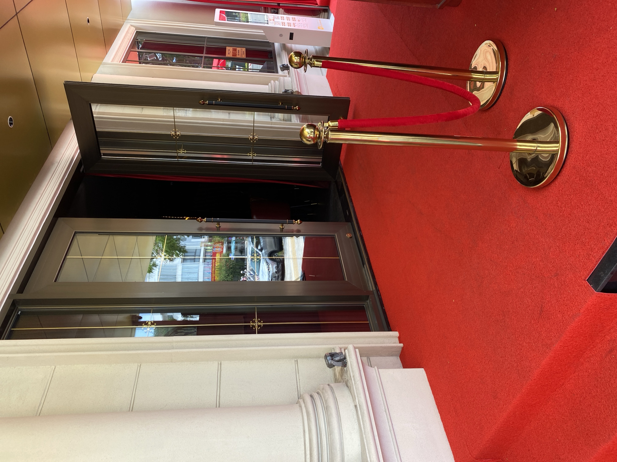 The image shows a red carpet leading to the entrance of a building with glass doors. There are stanchions with red velvet ropes on the carpet. The building has white columns and a gold-colored ceiling. In the reflection of the glass doors, a car and buildings can be seen. The scene appears to be set up for a special event or VIP entrance. Based on the visual cues, the location is likely a hotel, casino, or entertainment venue in Đà Nẵng, Vietnam. There are no people visible in the image.