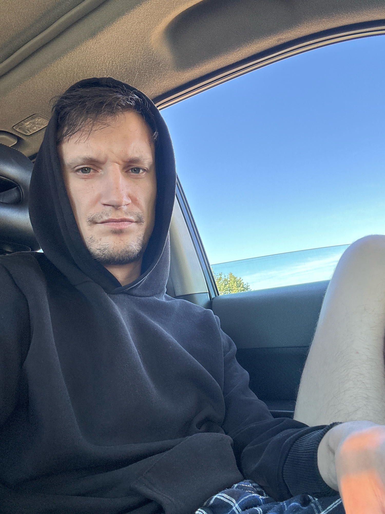 The image shows a man sitting in the passenger seat of a car, looking directly at the camera. He is wearing a black hooded sweatshirt with the hood up. His left leg is visible, wearing plaid shorts. Outside the car, a bright blue sky and trees are visible. The interior of the car appears to be dark, with a portion of the dashboard and ceiling visible. There is no information in the image to indicate the location in the city of Makiv, Ukraine.