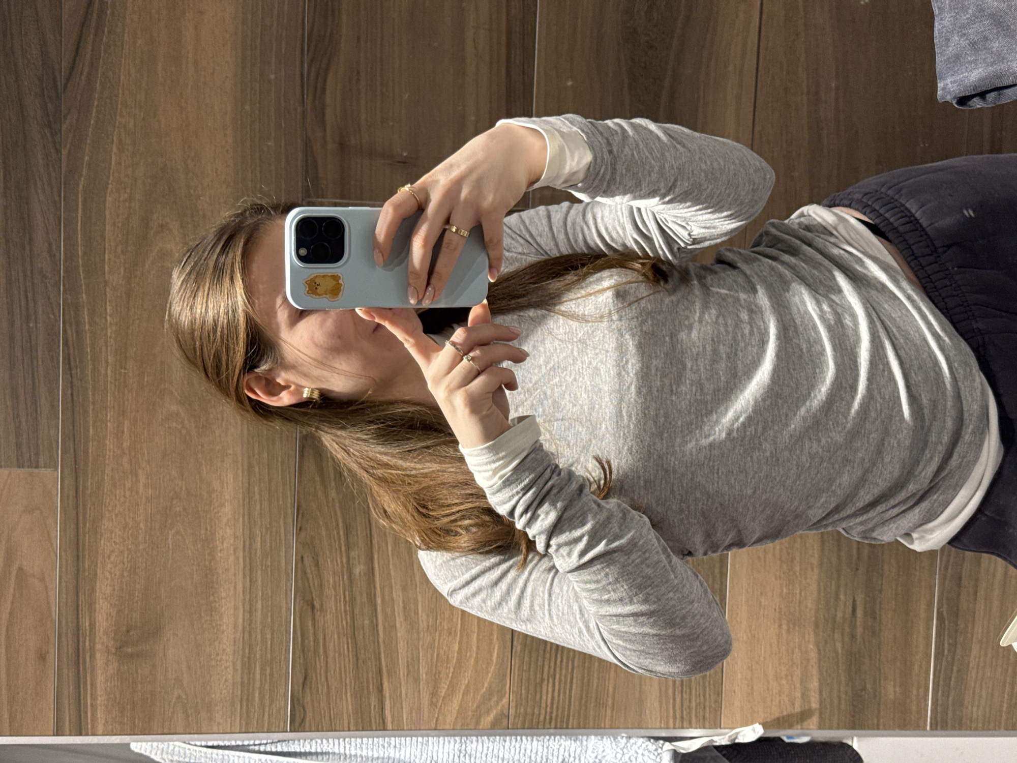 A person with long brown hair is taking a selfie in front of a wood-paneled wall or floor. The person is wearing a gray long-sleeved shirt with white cuffs and dark gray sweatpants. They are holding a light blue smartphone with a brown bear sticker on the back to take the photo. The person is wearing multiple gold rings on their fingers. It is not possible to determine the location is Khimki, Russia.