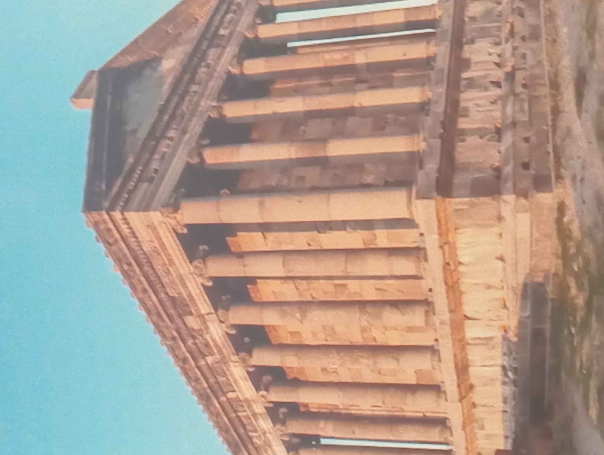 The image shows a low-angle, close-up view of an ancient architectural structure, possibly a temple or building ruin. The structure is built of stacked stone blocks, forming a series of horizontal layers and columns. The stones are a weathered, light brown color, suggesting age and exposure to the elements. The sky is a clear blue, visible in the background to the left of the structure. To the right of the structure is vegetation, possibly grass, with the ground appearing uneven.
The location of the structure is not discernible from the image, and it's impossible to state with certainty that it is in Karaganda, Kazakhstan, without additional information or context.