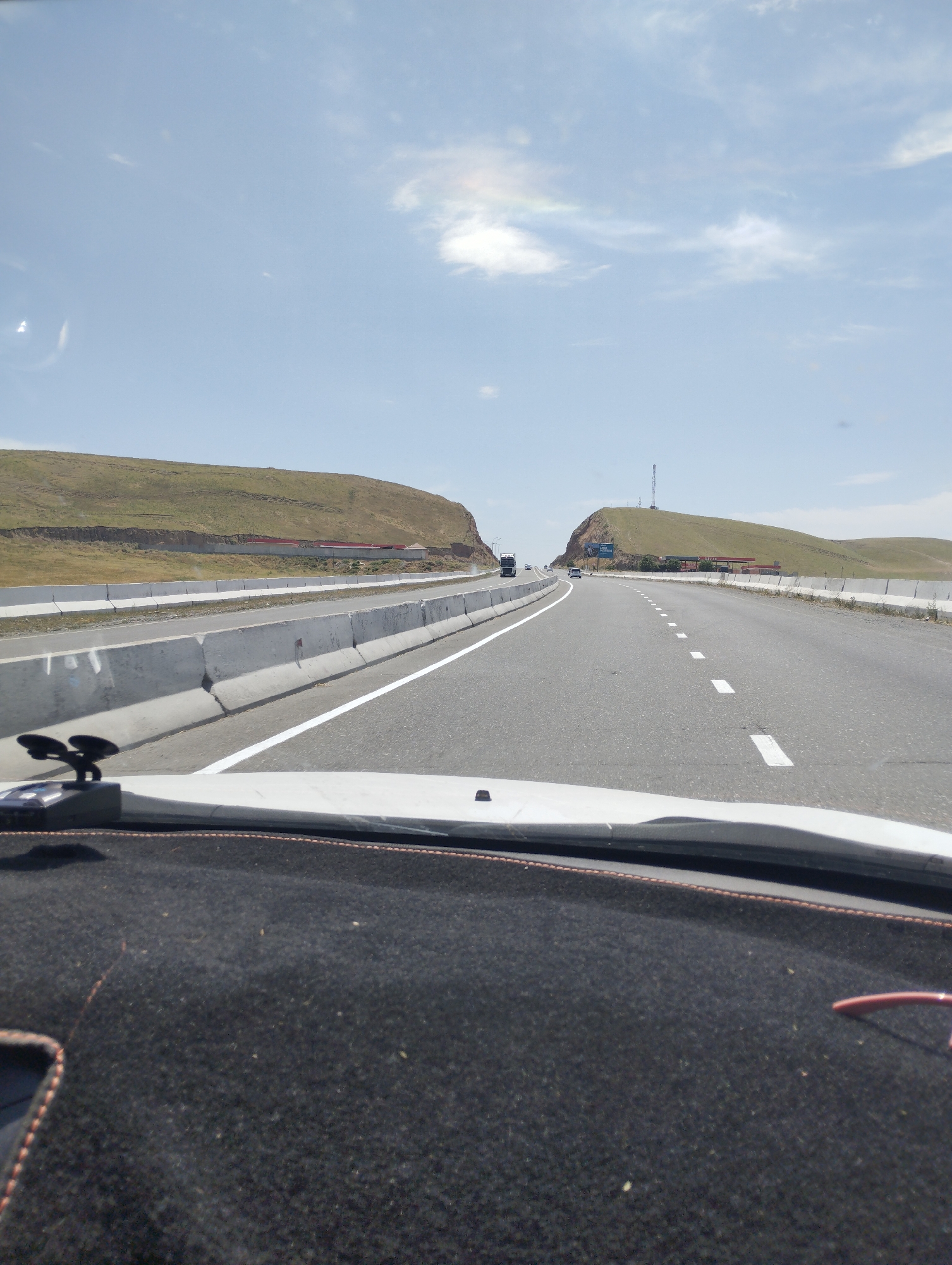 The image captures a view from inside a vehicle driving on a highway, presumably in Sangzar, Uzbekistan. The road is paved and has white lane markings. Concrete barriers line the sides of the road. In the distance, there are rolling hills covered with dry grass. The sky is blue with scattered clouds. A telecommunication tower is visible on one of the hills. The dashboard of the vehicle is visible in the foreground, with a black fabric cover and a small black device with suction cups. The overall scene depicts a daytime view of a highway landscape.
