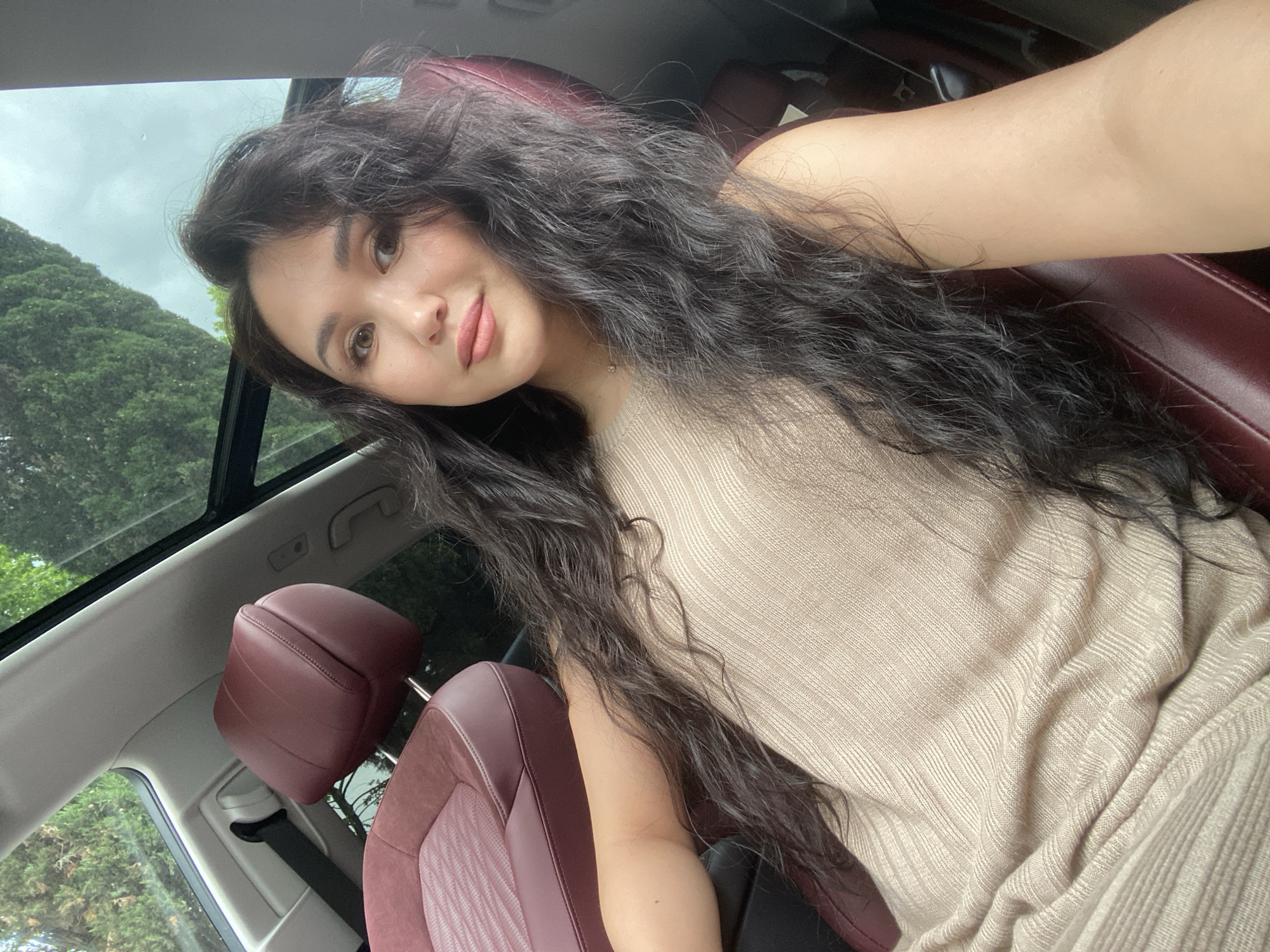 A young woman with long, dark, wavy hair is taking a selfie inside a car with a sunroof. She is wearing a light brown or beige garment. The car's interior features red leather seats. Through the sunroof, green trees and an overcast sky are visible. There is no indication of the location being Sochi, Russia.