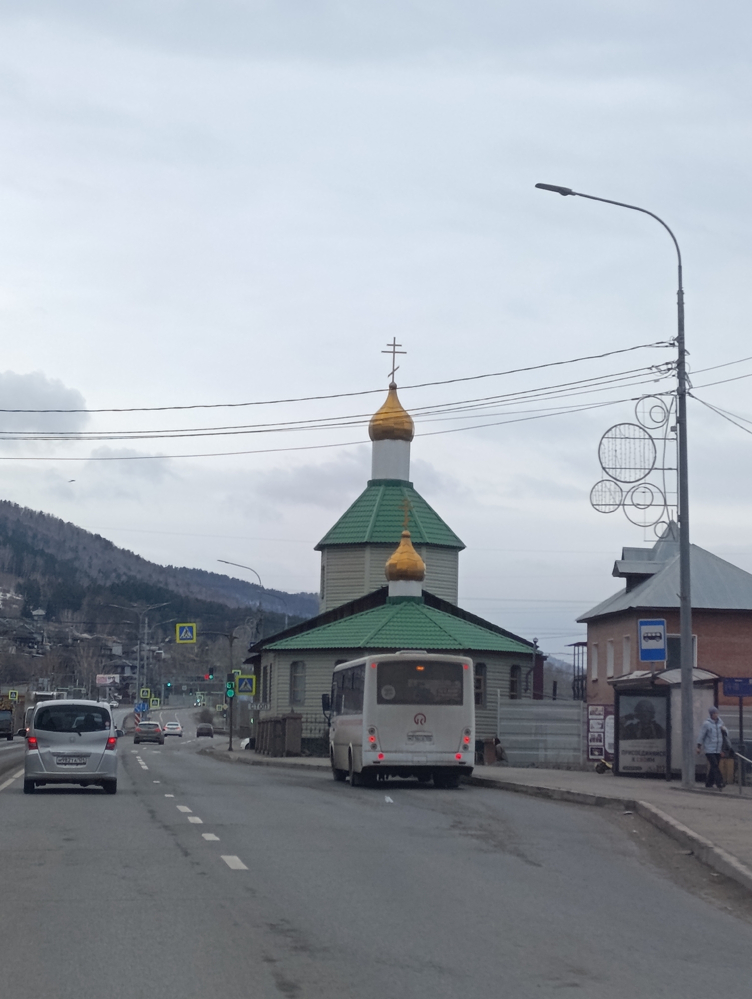 The image captures a street scene in Krasnoyarsk, Russia. In the center, there is a church with a green roof and two golden domes topped with crosses. A white bus is driving on the road in front of the church.
To the left, a silver minivan is driving in the same direction as the bus. Further down the road, other cars are visible. Pedestrian crosswalk signs are on both sides of the road. A mountain range is in the background on the left side.
On the right side, a bus stop is located on the sidewalk with a person standing nearby. A decorative streetlight is positioned next to the bus stop. Buildings of various architectural styles line the street. Overhead wires are strung across the sky. The sky is overcast.