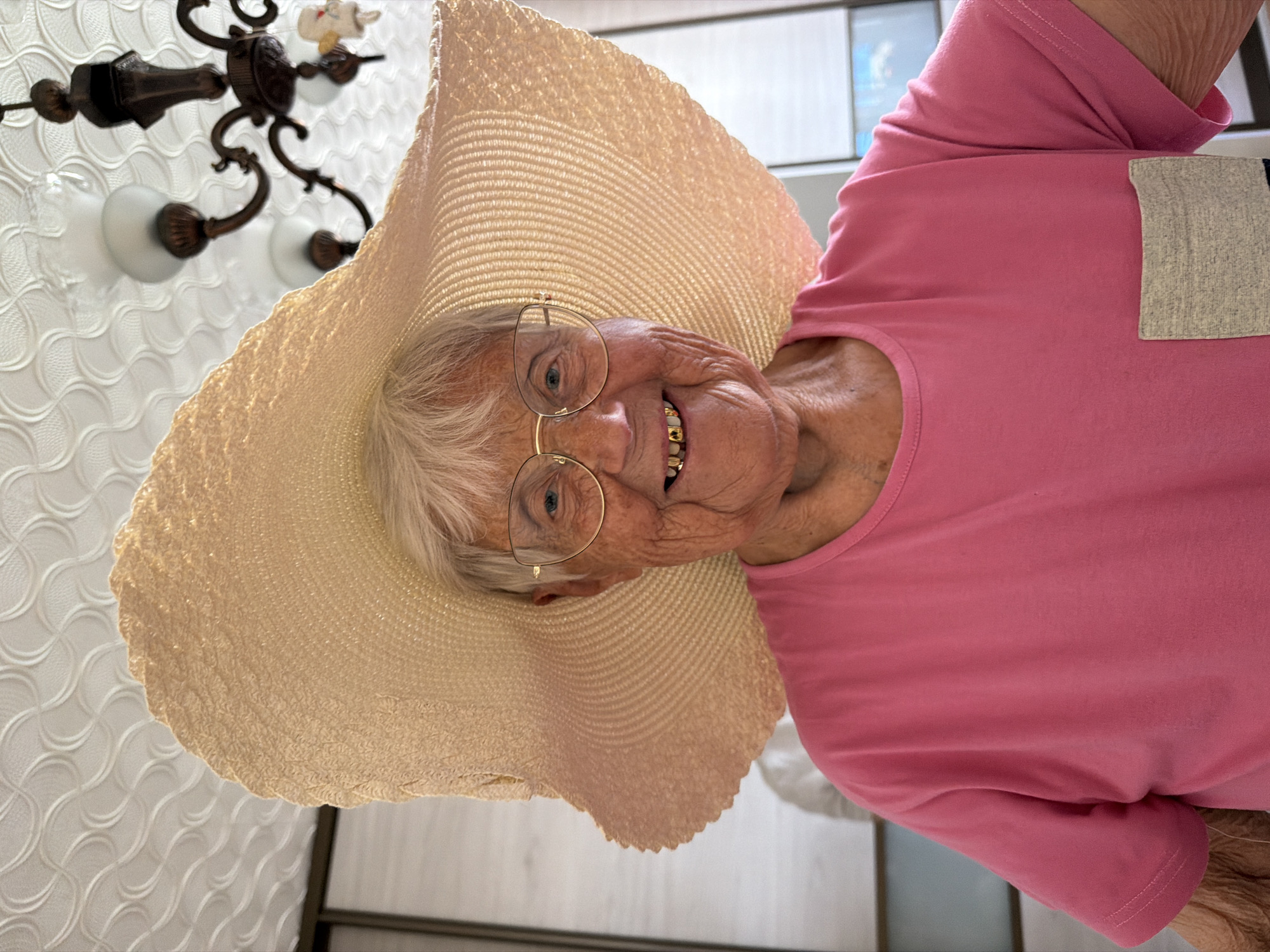 An elderly woman wearing a large straw hat and round gold-rimmed glasses smiles at the camera. She is wearing a pink shirt with a grey pocket. A chandelier is visible in the background. The location is Valozhyn, Belarus.