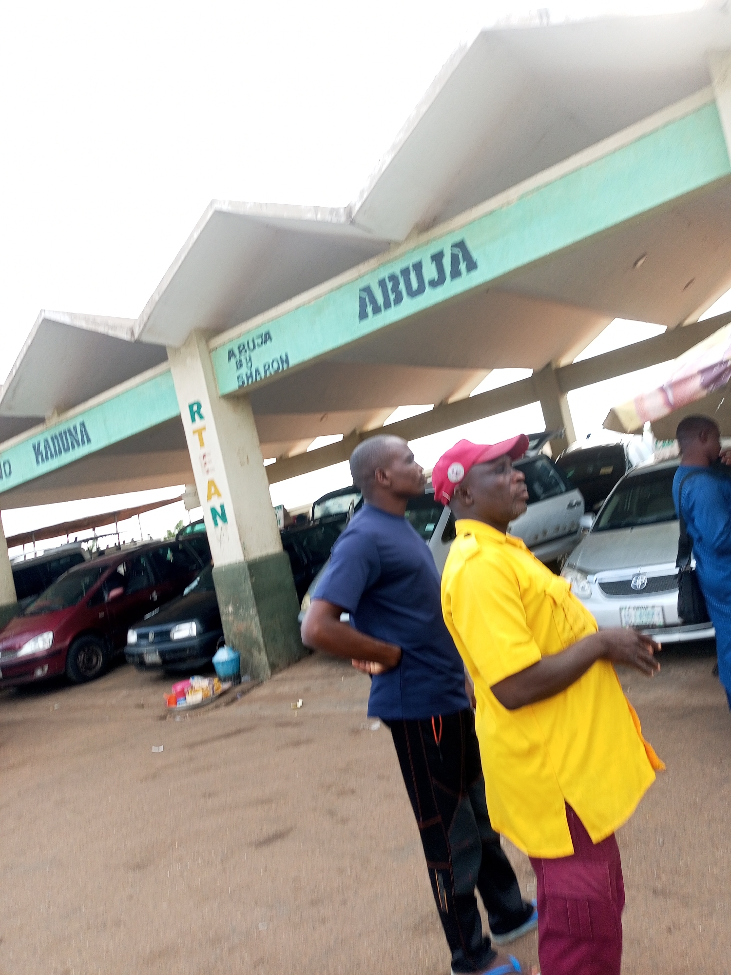 This media file captures an outdoor scene at what appears to be a transportation hub or bus station in Gombe, Nigeria. The primary subjects are three men standing under a covered structure, with several cars parked in the background. The men are positioned facing left in the frame, with their profiles visible. One man, in the foreground and to the right, wears a bright yellow shirt and a red baseball cap. He is gesturing with his hands. To his left, another man in a dark blue shirt and dark pants is also looking towards the left. A third man in a blue outfit is partially visible on the far right.
The setting is characterized by a concrete structure with a light-colored roof and turquoise-colored beams. The ground is a dusty, sandy surface. Several vehicles, including a red car and a dark-colored sedan, are parked under the shelter. In the background, other vehicles and structures are visible, suggesting a busy area.
The visible text on the turquoise beams reads "KADUNA" on the left side and "ABUJA" on the right side, indicating potential destinations or routes served by this location. There is also vertical text on a pillar that appears to read "RTEAN", which is likely an acronym related to transportation in Nigeria.
The lighting suggests it is daytime, with diffused light, possibly indicating an overcast sky or early morning/late afternoon. The overall atmosphere appears casual, with no overt signs of distress or urgency from the individuals. The scene suggests a moment of waiting or conversation at a travel point.