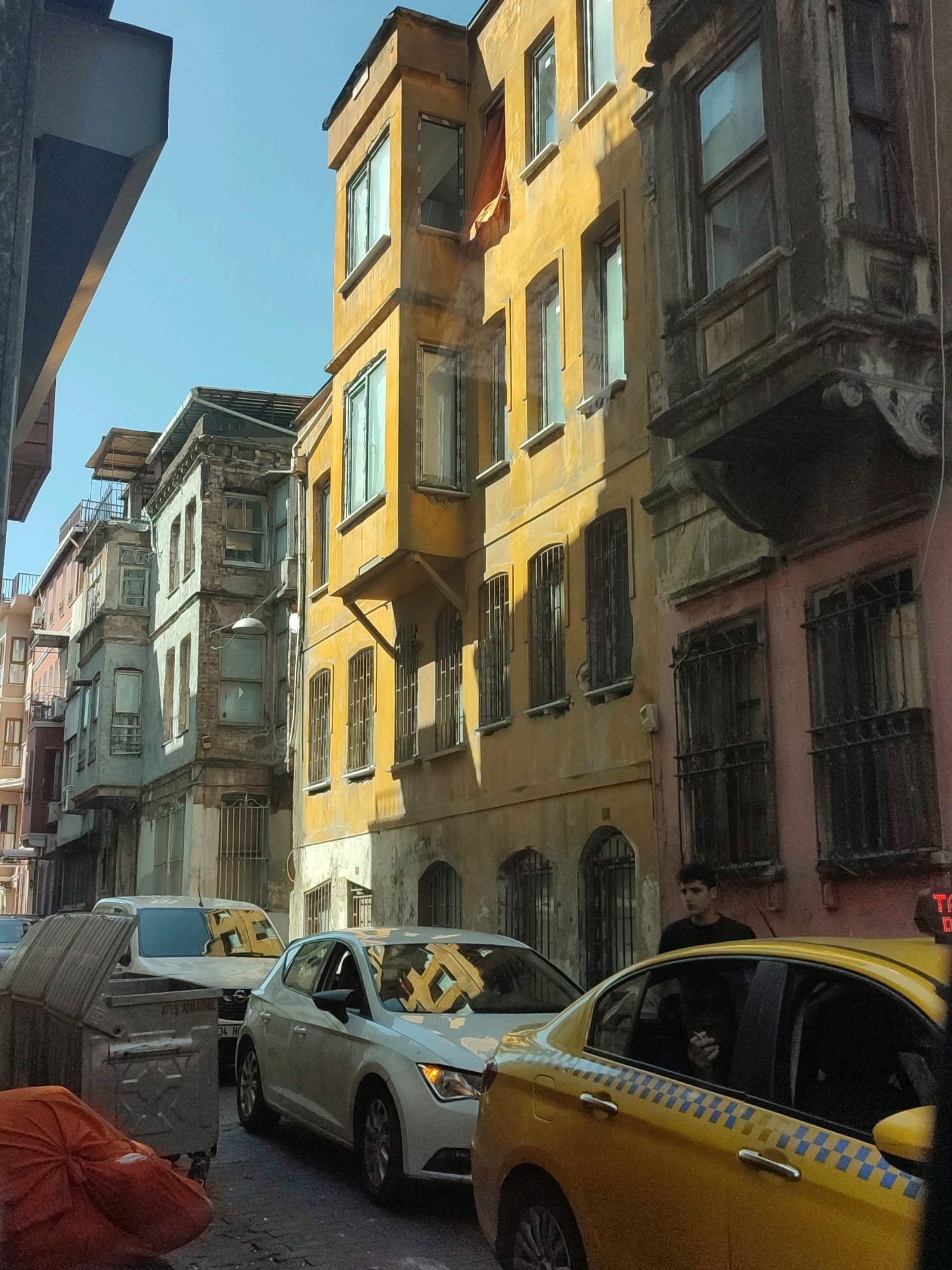 A sunny daytime street scene in Istanbul, Turkey, depicts a narrow alleyway flanked by old, weathered buildings of varying colors. The architecture is diverse, with some structures featuring ornate balconies and barred windows, while others are more plain. The buildings' facades range from faded yellow and pink to gray, suggesting age and exposure to the elements. A bright yellow taxi with blue checkered accents is prominently positioned in the foreground, its rear section visible. A white sedan is parked behind it, followed by another white vehicle, possibly a van. To the left, a large, grey dumpster with a hinged lid stands by the roadside, partially obscured by a crumpled orange tarp. A young man with dark curly hair is seen standing near the yellow taxi, looking towards the street. The sky is clear and blue.