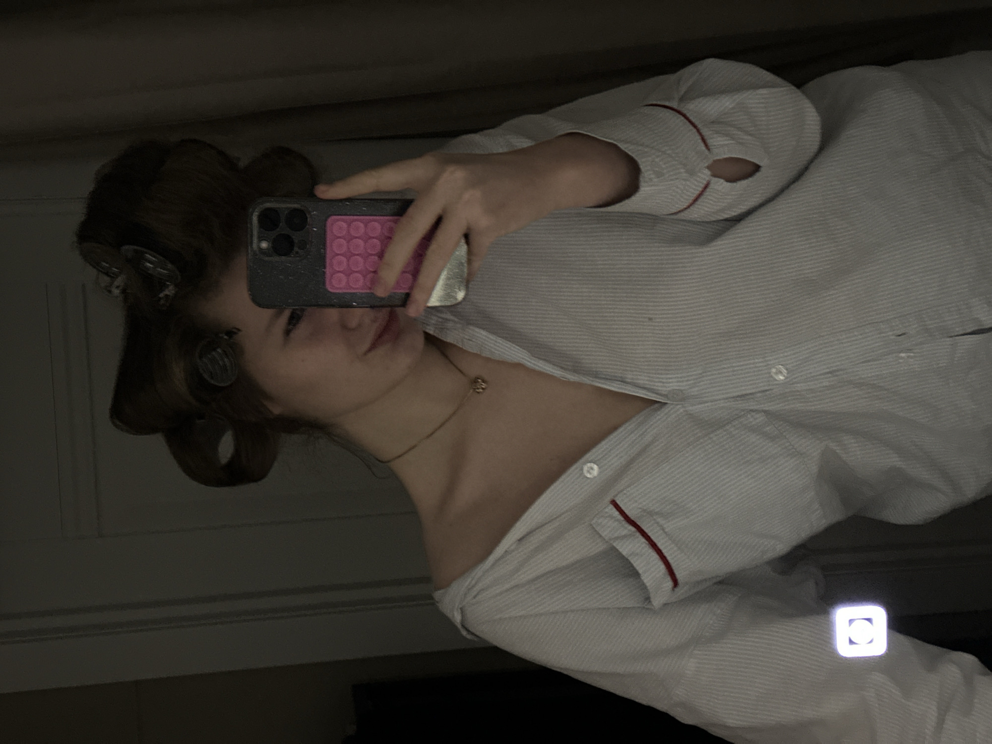 A person with hair rollers is taking a selfie in a dimly lit room. The person is wearing a striped pajama top with red trim and a delicate gold necklace. They are holding a smartphone with a dark-colored case and a pink, patterned design on the back. The background is dark and partially obscured, revealing a wall with molding and potentially a window. The location and city in Russia are not discernible from the image.