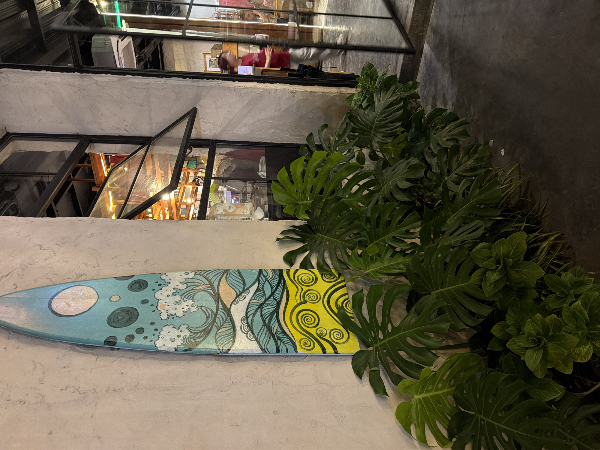 The image shows the exterior of a two-story building in Đà Nẵng, Vietnam. The building has a rough, textured white facade. A painted surfboard is mounted horizontally on the wall, featuring a design of waves and yellow swirls. To the right of the surfboard, there is a large cluster of Monstera Deliciosa plants. The building has metal-framed windows. The bottom window is open, revealing a glimpse of the interior with wooden shelves and assorted objects. The top window is reflecting the building across the street; a person wearing a red shirt is visible. The environment is urban, with a concrete walkway visible on the right side of the frame. The lighting suggests it is evening or night.