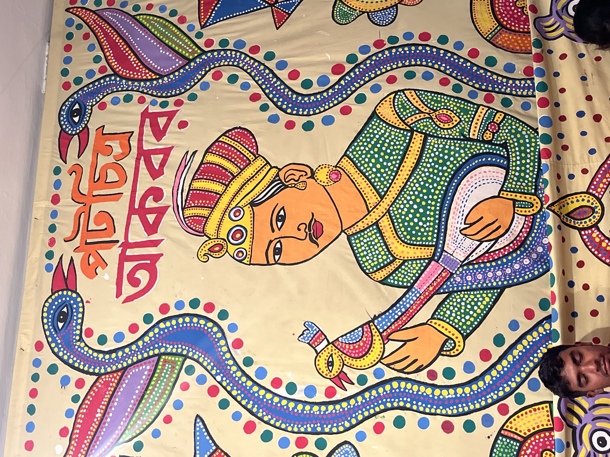 The image shows a hand-painted mural in Dhaka, Bangladesh. The artwork features a stylized man in traditional clothing and headwear, holding a bird. The artwork also shows decorated birds, Bengali text, and dotted patterns. There is an individual visible at the right edge of the image.