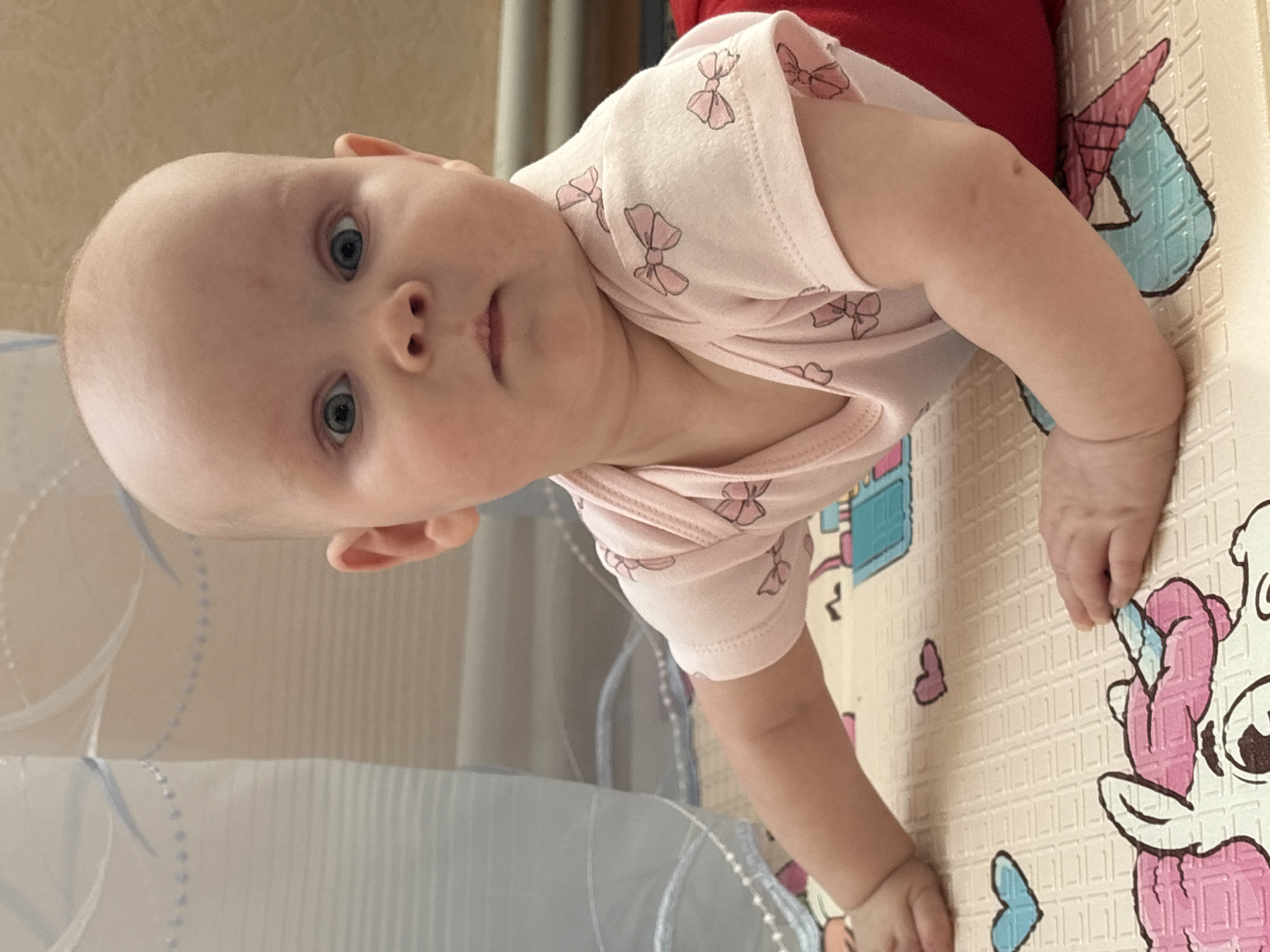A baby with blue eyes is lying on a play mat. The baby is wearing a light pink onesie with a bow pattern and red pants. The baby is fair-skinned. The play mat is light beige with various colorful cartoon figures. A sheer, blue-trimmed curtain is visible in the lower-left corner. The location is Votkinsk, Russia.