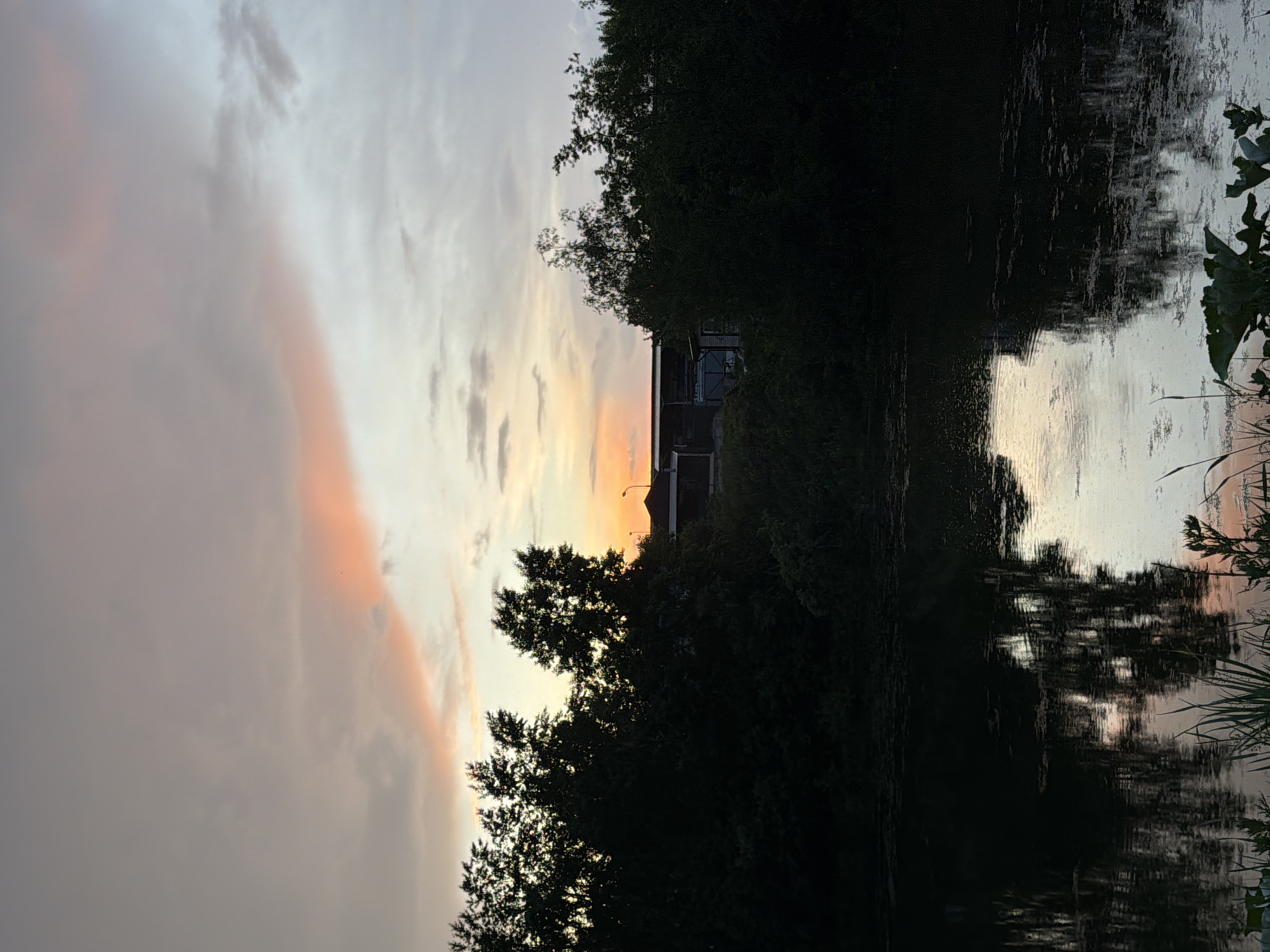 The image captures a serene landscape scene during either sunrise or sunset, set in Chaikovsky, Russia. The sky, occupying the upper portion of the frame, displays a soft gradient of colors, transitioning from light grey and pale blue at the top to warm orange and pink hues closer to the horizon. A prominent, elongated cloud band of vibrant orange runs horizontally across the mid-upper sky.
Below the colorful sky, a dense line of trees forms a dark silhouette, obscuring the horizon. Through and above this treeline, a dark, angular building, possibly an industrial structure or a large shed, is partially visible. Its dark form is sharply contrasted against the brighter sky behind it.
The lower part of the image features a body of water, likely a river or canal, which beautifully reflects the sky's soft colors and the dark silhouettes of the trees and the building. The reflections are slightly rippled, adding depth and texture to the scene. Sparse reeds or aquatic plants can be seen along the water's edge on the right. The overall atmosphere is one of peaceful twilight, with no discernible human activity or visible text.