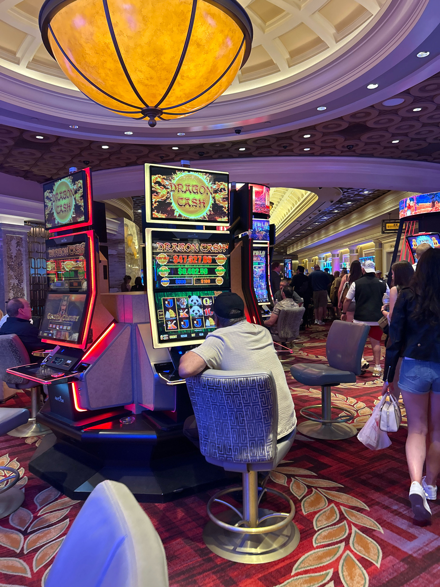 The image depicts the interior of a casino in Paradise, Nevada, United States of America. The scene is dominated by rows of brightly lit slot machines, with "Dragon Cash" branding prominently displayed on several of them. In the foreground, a man is seated at a slot machine, facing away from the camera, with his back towards the viewer. To his right, another slot machine is visible, and a woman in denim shorts and a black jacket walks away from the camera, carrying a white handbag. The casino floor is carpeted with a red and gold leaf pattern. Overhead, a large, ornate yellow light fixture hangs from the ceiling, and the ceiling itself features patterned tiles and recessed lighting. Other casino patrons are visible in the background, walking through the aisles between the slot machines. The overall atmosphere suggests a lively and active gambling environment.