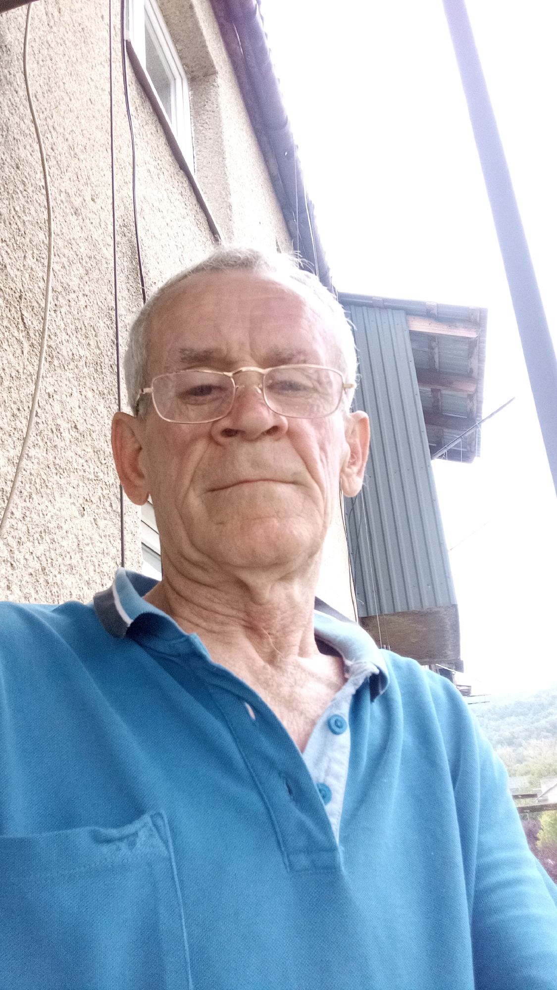 A man with fair skin and grey hair wears glasses and a blue polo shirt. He is outdoors, with a textured wall and a window visible behind him. A corrugated metal structure and some greenery are also in the background. The overall lighting suggests it is daytime.