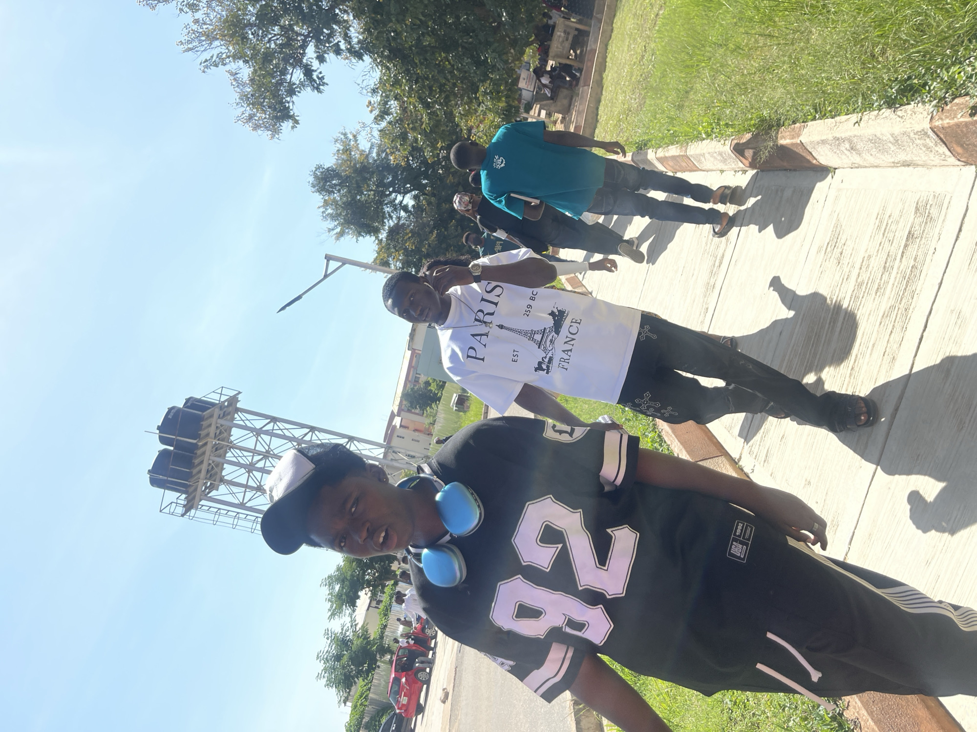 The image captures a sunny day on a paved walkway in Rafin Par, Nigeria. In the foreground, a young person wearing a black jersey with white lettering and blue headphones around their neck is walking towards the camera. To the right, another young person is walking away, wearing a white t-shirt with "PARIS EST 259 BC FRANCE" printed on it, and dark pants. Several other individuals are also visible walking on the path behind them. The environment includes lush green grass bordering the walkway, trees, and buildings in the background. A large industrial water tower stands tall against a clear blue sky. A red car is parked in the distance. The scene suggests a casual outdoor setting, possibly on a university campus or in a public area.