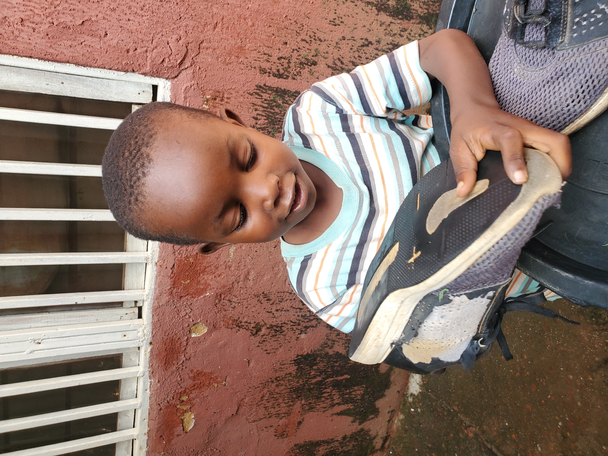A young Black child is shown from a high angle, sitting with their eyes closed and a gentle smile on their face. They are wearing a striped t-shirt with white, blue, black, and orange stripes. The child is holding a pair of well-worn black and gray athletic shoes. One shoe is particularly tattered, with significant wear and tear on the sole and toe.
The child is seated next to a textured, reddish-brown wall with visible signs of weathering and some peeling paint. To the left of the child, there is a white-barred window or grate set into the wall. The ground beneath and around the child appears to be damp and possibly muddy, with patches of green moss or algae.
The scene suggests an outdoor setting, possibly a residential area in Jos, Nigeria. The lighting is soft, indicating it could be daytime, but the specific time of day or weather conditions are not clearly discernible. The child's expression conveys a sense of contentment or peace. There is no visible text in the image.
