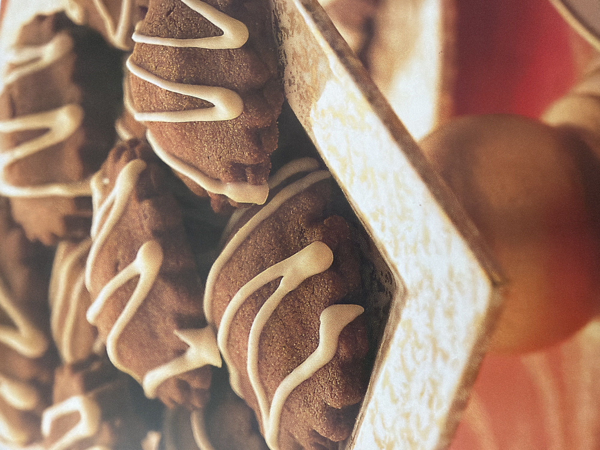 The image shows a close-up of several chocolate cookies, possibly pastries, arranged in a decorative box. The cookies are dark brown and appear to have a textured surface, possibly from nuts or spices. They are drizzled with a white icing in a decorative pattern. One cookie in the foreground is clearly visible with its intricate icing design. The box itself is light-colored, possibly wood or cardboard, with a textured or embossed pattern on its inner surface. In the background, out of focus, is a soft, warm-toned area, which could be part of a tablecloth or another decorative element. The overall lighting is warm and inviting, suggesting a cozy or festive setting, perhaps a bakery, a home kitchen, or a gift presentation. There are no people visible in the image, and no text is discernible. The focus is entirely on the appetizing appearance of the cookies and their presentation. The shallow depth of field blurs the background, drawing attention to the detailed textures and patterns of the cookies and the box.