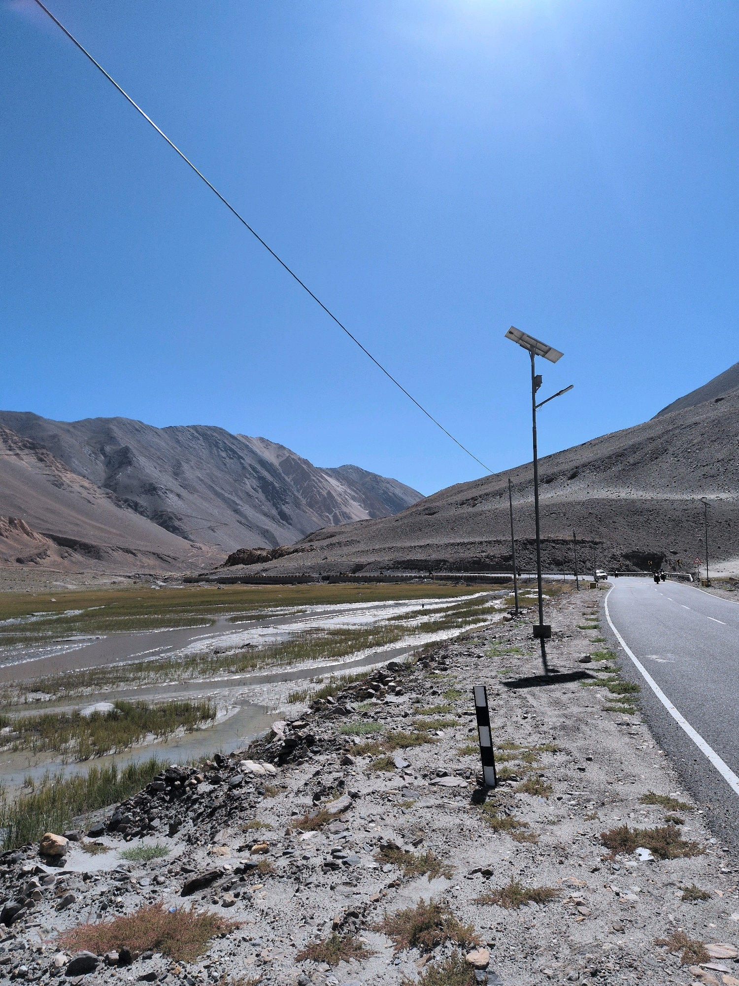 The image depicts a mountainous, arid landscape under a clear blue sky. A paved road curves along the right side of the frame, with a rocky, sparse terrain bordering it. A single-pole street light with a solar panel is prominently positioned near the road. In the distance, a wide, shallow body of water with sparse vegetation stretches across a valley, framed by barren, grey mountains. A power line is visible diagonally across the sky. The scene appears to be a remote, high-altitude location, possibly in Ladakh, India, given the typical terrain and presence of solar-powered street lights common in such regions.