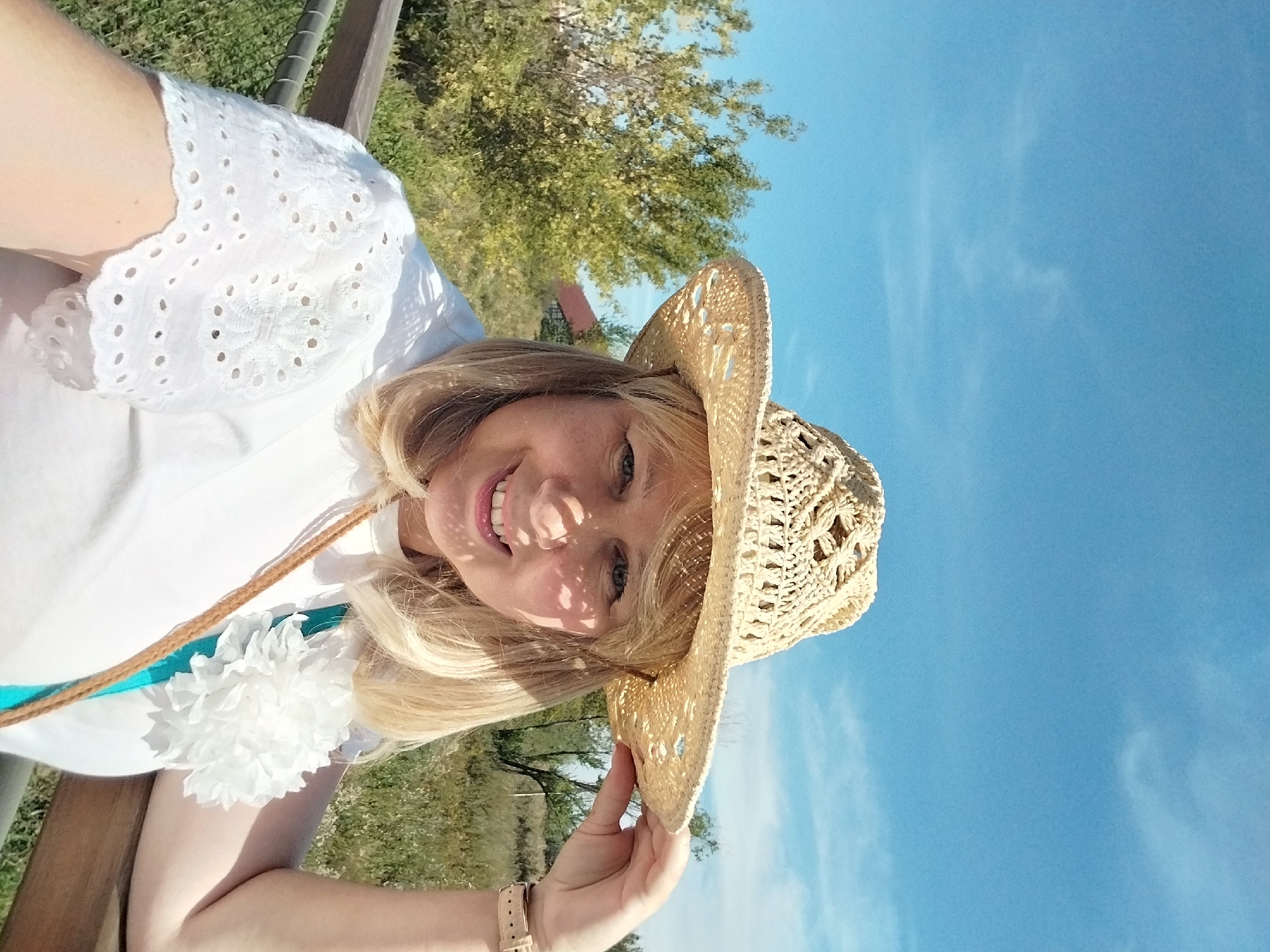 A woman wearing a straw hat and a white shirt with eyelet sleeves is captured in a selfie. She is holding the brim of her hat with her right hand. A light brown strap from a bag or purse is visible crossing her chest, and a white floral embellishment is attached to her shirt. She has blonde hair and is smiling. The background shows trees and vegetation under a clear blue sky with some wispy clouds. The scene appears to be outdoors, possibly in a park or natural area, and given the context of Orenburg, Russia, this could be a recreational area within the city.
