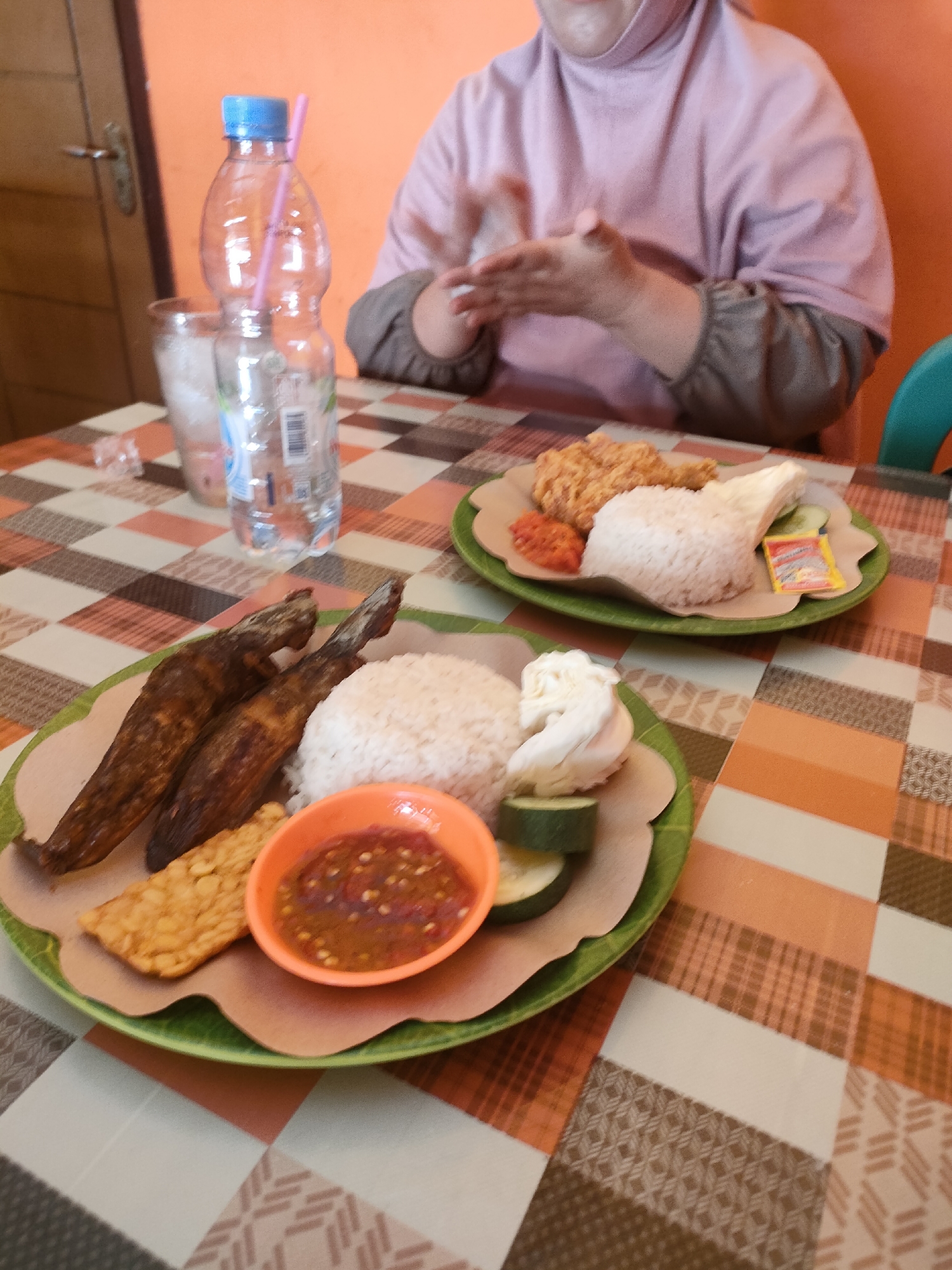 The image depicts two plates of Indonesian food, likely served in a casual dining setting in Pringsewu, Indonesia. Both plates contain rice, fried items, and accompaniments. The foreground plate features two pieces of fried fish, a slice of fried tempeh, a small bowl of sambal (chili sauce), and sliced cucumber. The plate behind it shows fried chicken, rice, sliced cucumber, and a small packet of condiment. A person wearing a pink hijab and a long-sleeved shirt is partially visible in the background, reaching for something on the table. A plastic bottle of water with a blue cap and a pink straw stands to the left, along with a glass of ice. The table is covered with a checkered tablecloth in shades of orange, brown, and white. The background wall is a uniform orange color. The overall impression is of a meal being enjoyed in a local eatery.