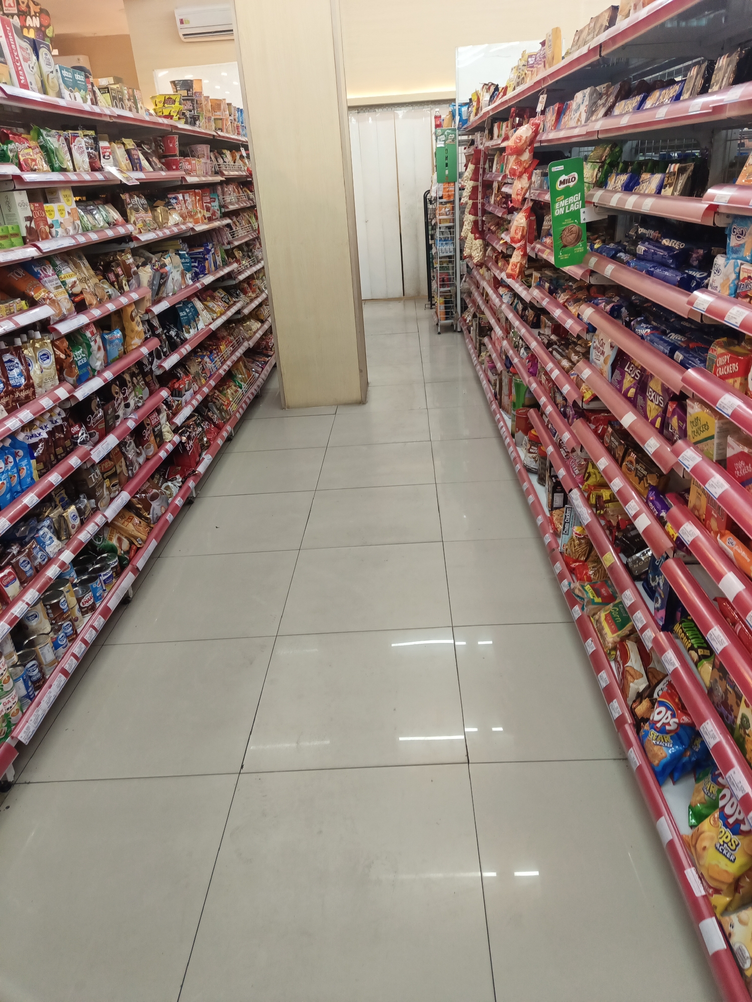 The image depicts the interior of a convenience store, likely a minimarket, in North Jakarta, Indonesia. The perspective is from the aisle between two long shelving units that are filled with various packaged food items, snacks, and drinks. The shelving units are painted red with white price tag holders. The floor is tiled in a light beige color. The lighting appears to be from overhead fluorescent lights, creating a bright and uniform illumination. The overall impression is one of a well-stocked and organized retail space.