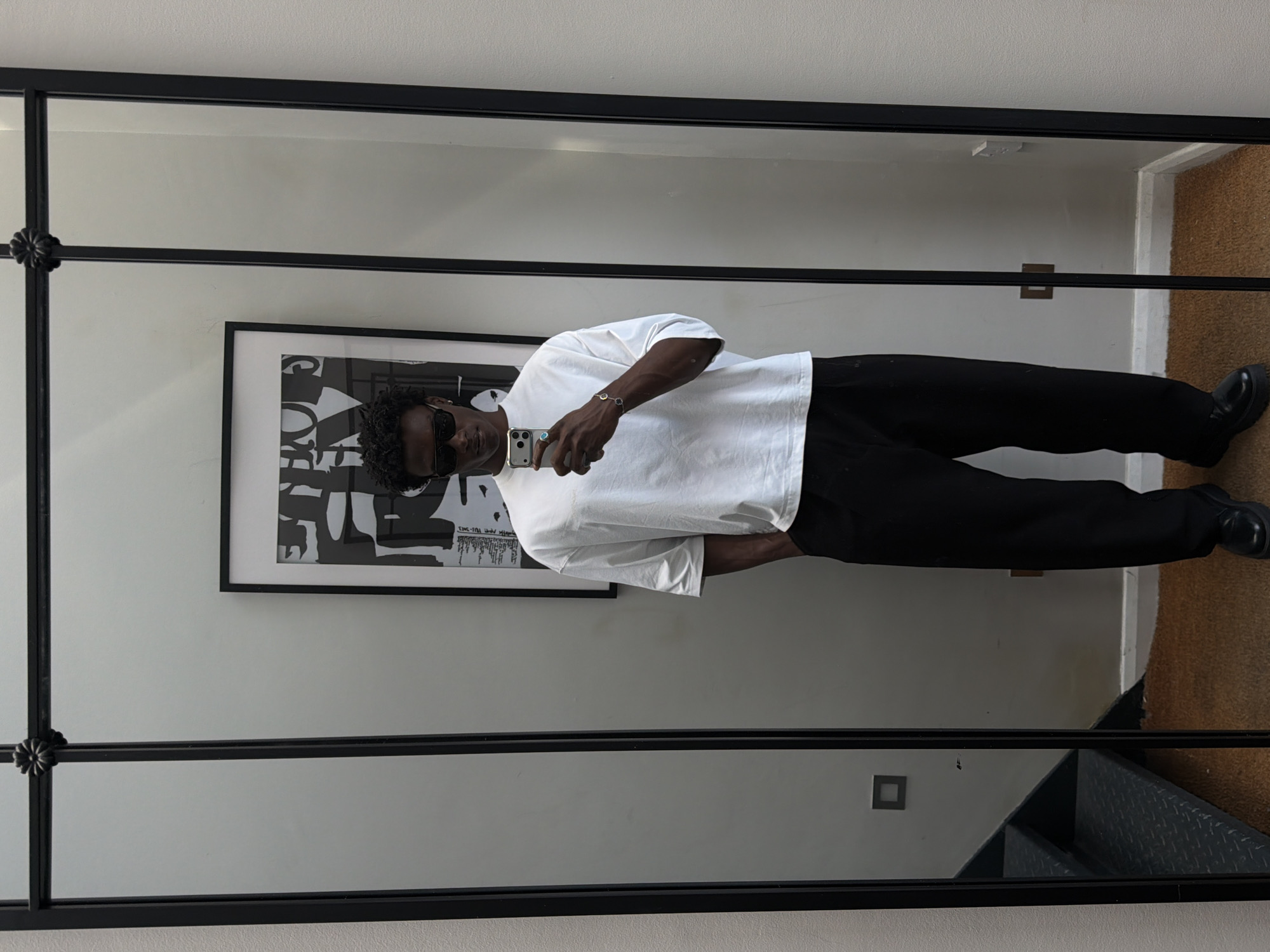 A full-length mirror reflects a person standing in what appears to be a room. The person, dressed in a white t-shirt and black trousers, is holding a smartphone and taking a mirror selfie. The background of the reflection shows a framed black and white artwork on the wall, a set of stairs in the corner, and a glimpse of the floor. The mirror itself has a black frame with decorative elements at the top. The environment appears to be indoors, possibly a home or a studio, and there is no indication of the location being in the city of London, United Kingdom.