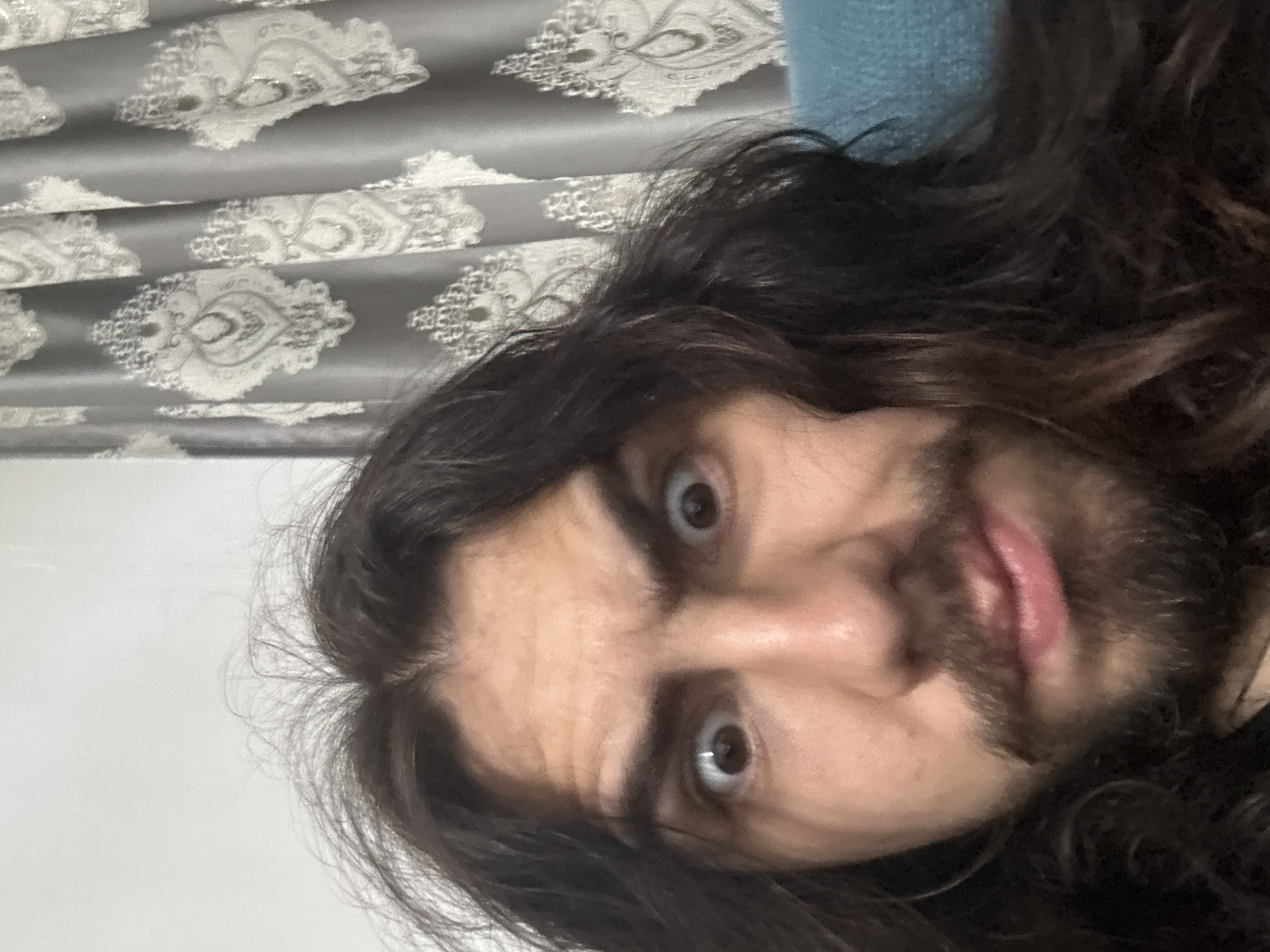 A close-up selfie shows a person with long, dark, unkempt hair and a mustache and beard. The person's eyes are wide and their eyebrows are raised, giving them a surprised or alarmed expression. They are wearing a dark shirt, and behind them, a gray curtain with an ornate pattern is visible. The background also includes a sliver of a teal-colored surface, possibly a couch. The image is taken indoors with soft lighting.
