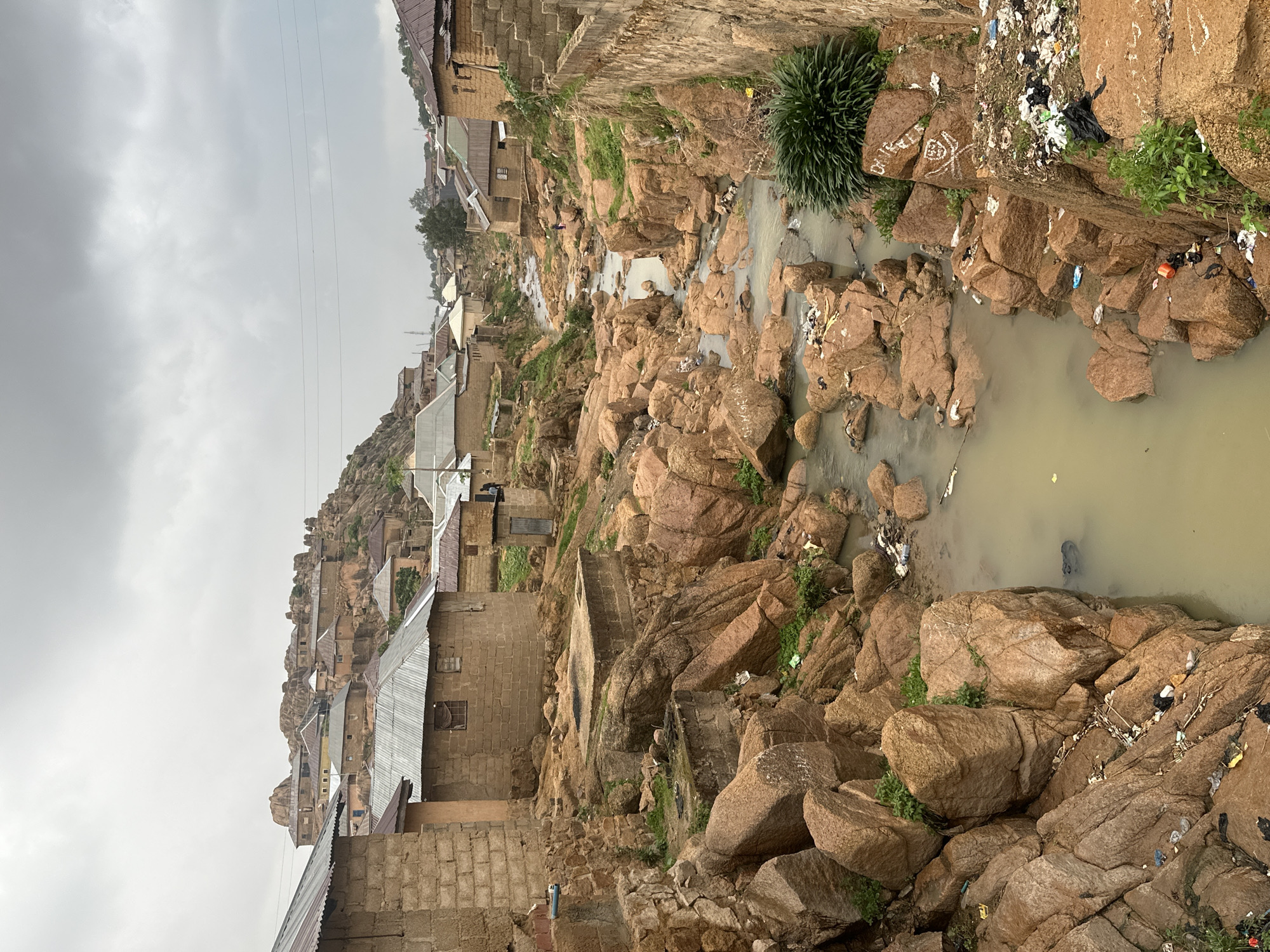 This media file captures an aerial-style view of a dense, informal settlement built on a rugged, rocky landscape, typical of the terrain found in places like Jos, Nigeria. The image is presented vertically (rotated 90 degrees clockwise).
The scene is dominated by numerous large, reddish-brown rock formations that form the natural foundation and pathways of the area. Amidst and atop these rocks, a multitude of low-rise buildings are clustered tightly. These structures are primarily constructed from concrete blocks and feature corrugated metal roofs, indicating a practical and often informal style of architecture.
A narrow, shallow stream of murky, yellowish-brown water winds its way through the rocky channel, acting as a central element of the settlement. A significant and noticeable detail is the widespread environmental pollution: substantial amounts of refuse, including plastic bags, bottles, and other debris, are scattered across the rocks along the stream banks and are visible floating in the water. Piles of trash are evident on various rock ledges and along the waterway.
Sparse green vegetation and small bushes grow in patches wherever soil is present among the rocks. The sky above is overcast and uniformly grey, indicating a cloudy or heavily subdued daytime. Power lines are visible stretching across the upper portion of the frame. No people or specific activities are discernibly taking place, suggesting a quiet moment. Some white, graffiti-like markings are present on the rocks; the word "Dunia" is faintly visible on one of them.