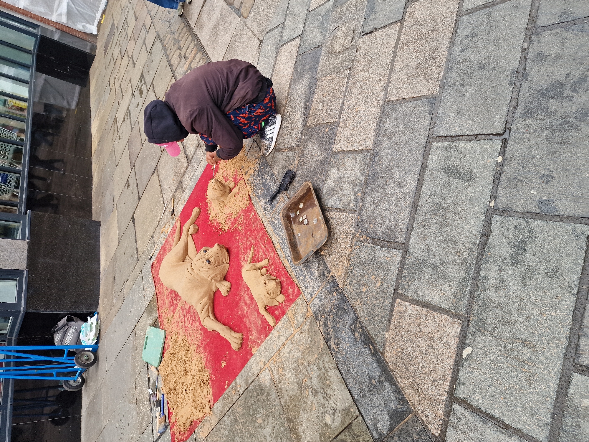 A street artist in Durham, United Kingdom, is creating sand sculptures of dogs on a red cloth placed on a paved public walkway. The artist, wearing a black hooded jacket with a pink hat and patterned trousers, is bending over the sculptures, making adjustments. Two dog sculptures are visible. A tray with coins, likely donations, is next to the artist, along with brushes and a trowel. A blue trolley cart and a building with glass windows are in the background. The walkway consists of light and dark grey paving stones.