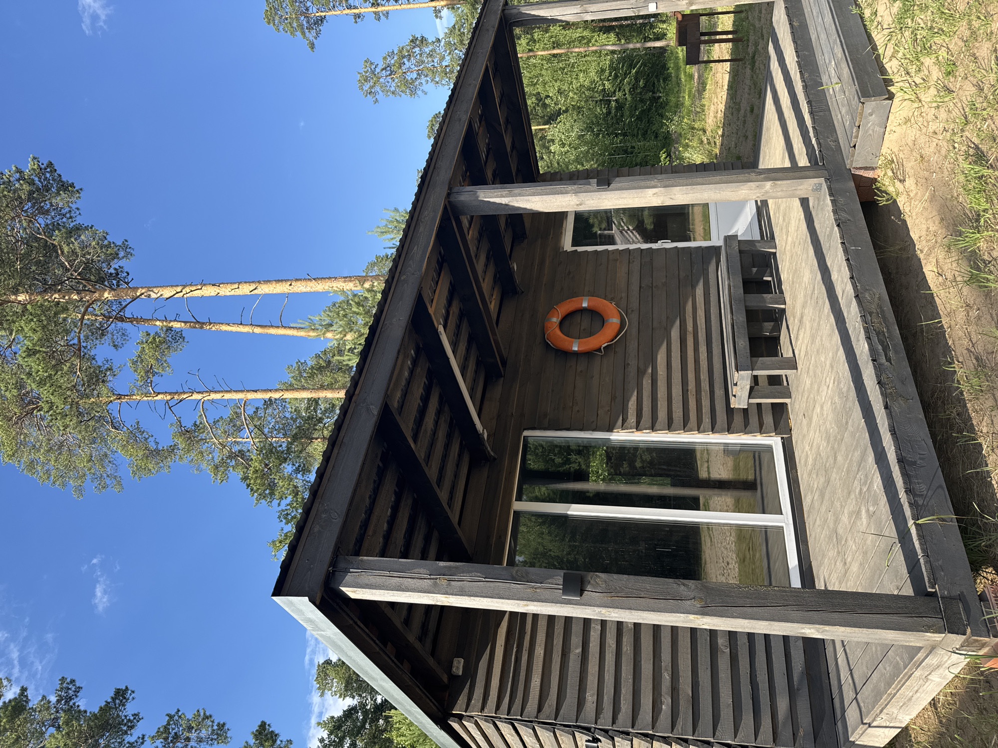 The image shows a dark wooden cabin with a covered porch in the city of Novyi, Russia. The cabin has large windows and a sliding glass door. A life preserver is hanging on the wall of the cabin. There is a wooden bench on the porch. In the background, there are tall pine trees and a clear blue sky. A grill is visible to the right of the cabin. The ground is sandy with some grass. The image appears to be taken in a natural, possibly rural or recreational setting.