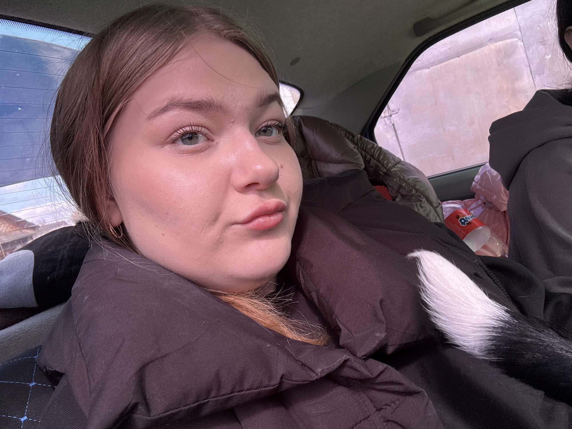 The image shows a young woman in a car, facing the camera with slightly pursed lips. She has light brown hair and blue eyes. She is wearing a dark, puffy jacket. Part of a black and white animal tail rests on her jacket. In the background, another person can be seen partially, wearing a dark hoodie. A brown, quilted jacket and a bottle of juice are also visible. The location is suggested as Узяр, Russia. The context implies a scene within the interior of a vehicle.