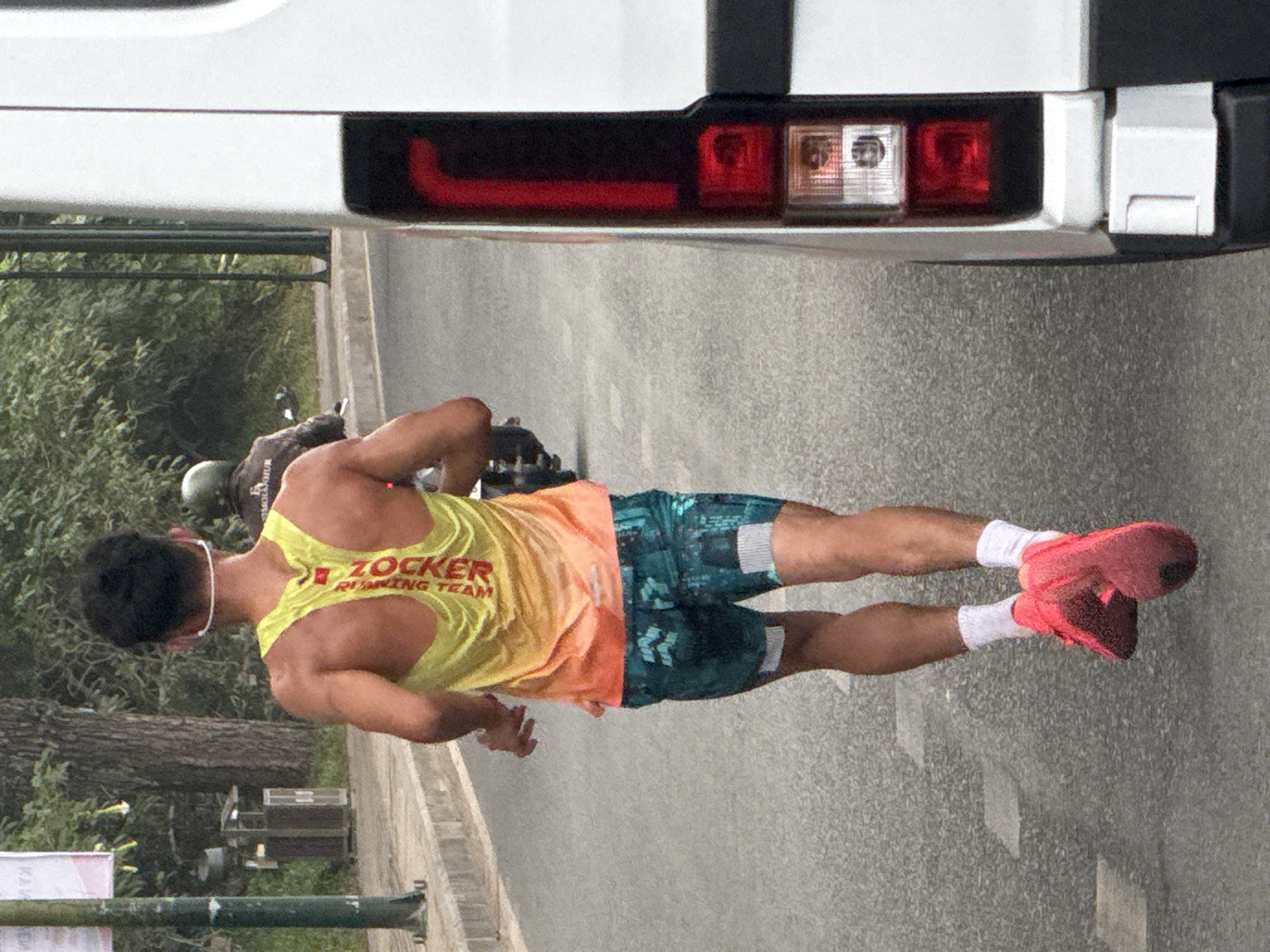 A man runs on a paved road in Da Lat, Vietnam, wearing a yellow and orange tank top with "ZOCKER" and "RUNNING TEAM" printed vertically on the back, and teal and black patterned shorts. He also wears white socks and bright pink running shoes. A white van is partially visible on the right side of the frame, and a motorcycle with a rider wearing a helmet and dark jacket is in the background on the left. Trees and foliage line the side of the road. A trash receptacle and a lamppost are also visible in the background.