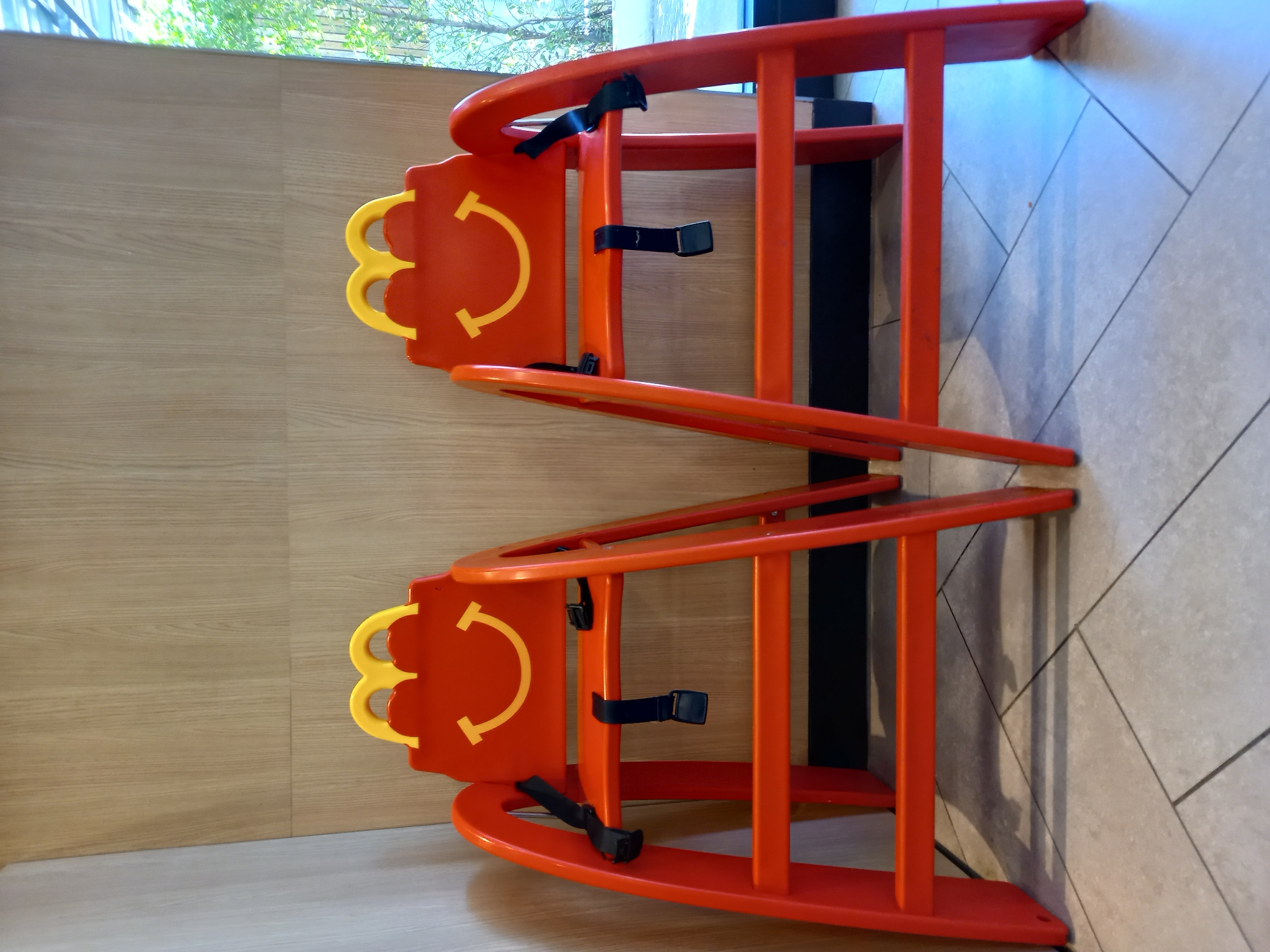 Two identical orange high chairs, each adorned with a yellow Happy Meal smile and eyebrows, are positioned side-by-side in a well-lit interior. The chairs have black safety straps. The background consists of a wood-paneled wall and a tiled floor. To the right, a window offers a view of green foliage and sky. The scene is likely within a restaurant or family-friendly establishment.