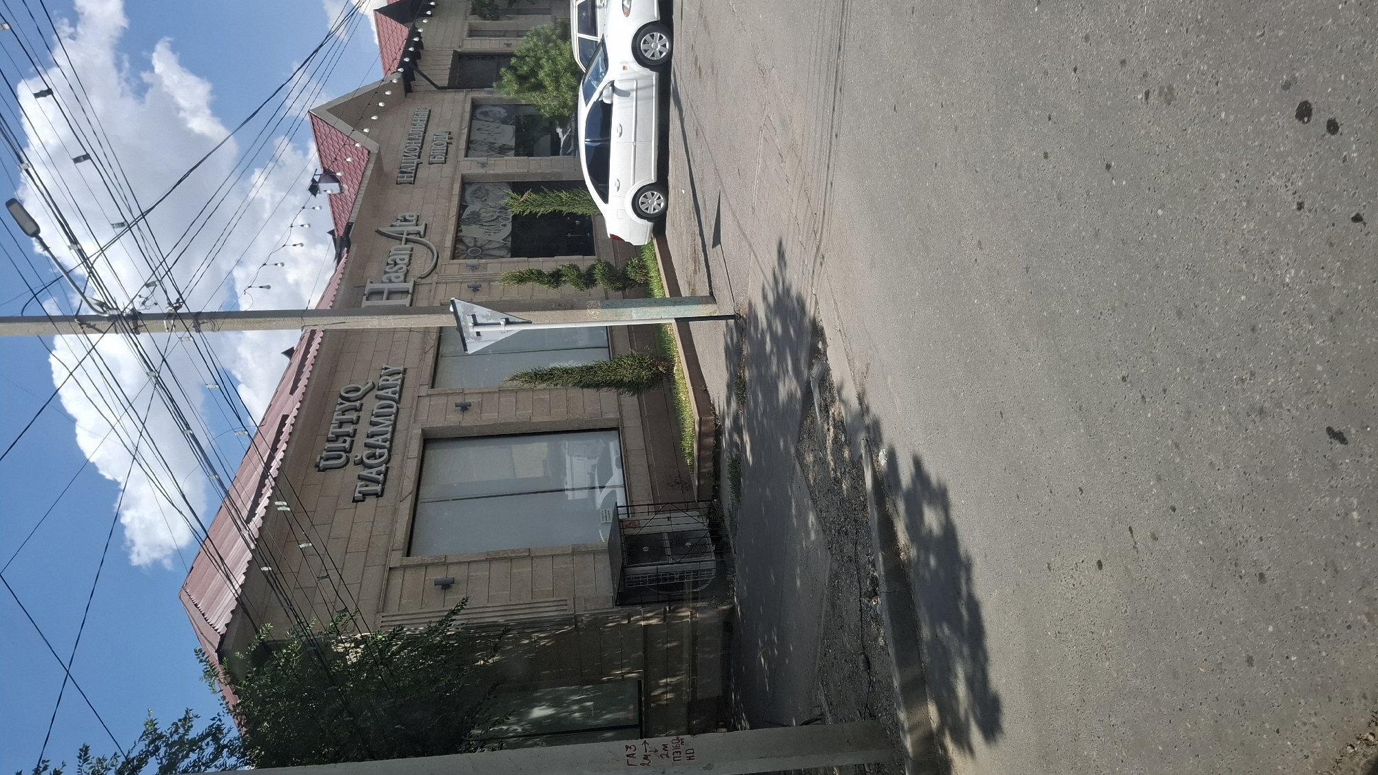 The image captures a street scene in Shymkent, Kazakhstan, on a bright, sunny day with scattered clouds. The primary focus is a light-colored, stone-clad building with a reddish-brown tiled roof. Two signs are visible on the building's facade. The larger sign, in silver lettering, reads "ULTTYQ TAGAMDARY." To its right, another sign, also in silver, displays "Hasan Ata" followed by "НАЦИОНАЛЬНЫИ БЛЮД". The building appears to house commercial establishments, indicated by large, reflective windows and what seems to be a restaurant or cafe.
In front of the building, a sidewalk separates it from the street. Several tall, slender trees are planted along the edge of the sidewalk. A white sedan is parked on the right side of the image, facing away from the viewer. Another white vehicle, partially visible, is parked behind it. A traffic sign, shaped like an inverted "T" with a horizontal bar, is mounted on a pole near the sidewalk.
To the left of the frame, a utility pole with numerous overhead wires is prominent. A metal fence encloses an outdoor air conditioning unit situated near the building's foundation. The street itself is paved and shows signs of wear, with some patches and markings. Shadows from trees and overhead wires are cast across the sidewalk and street, indicating the time of day. A small section of a concrete curb is visible at the edge of the street. On the left-hand utility pole, there are faint markings that appear to be graffiti or informational labels.