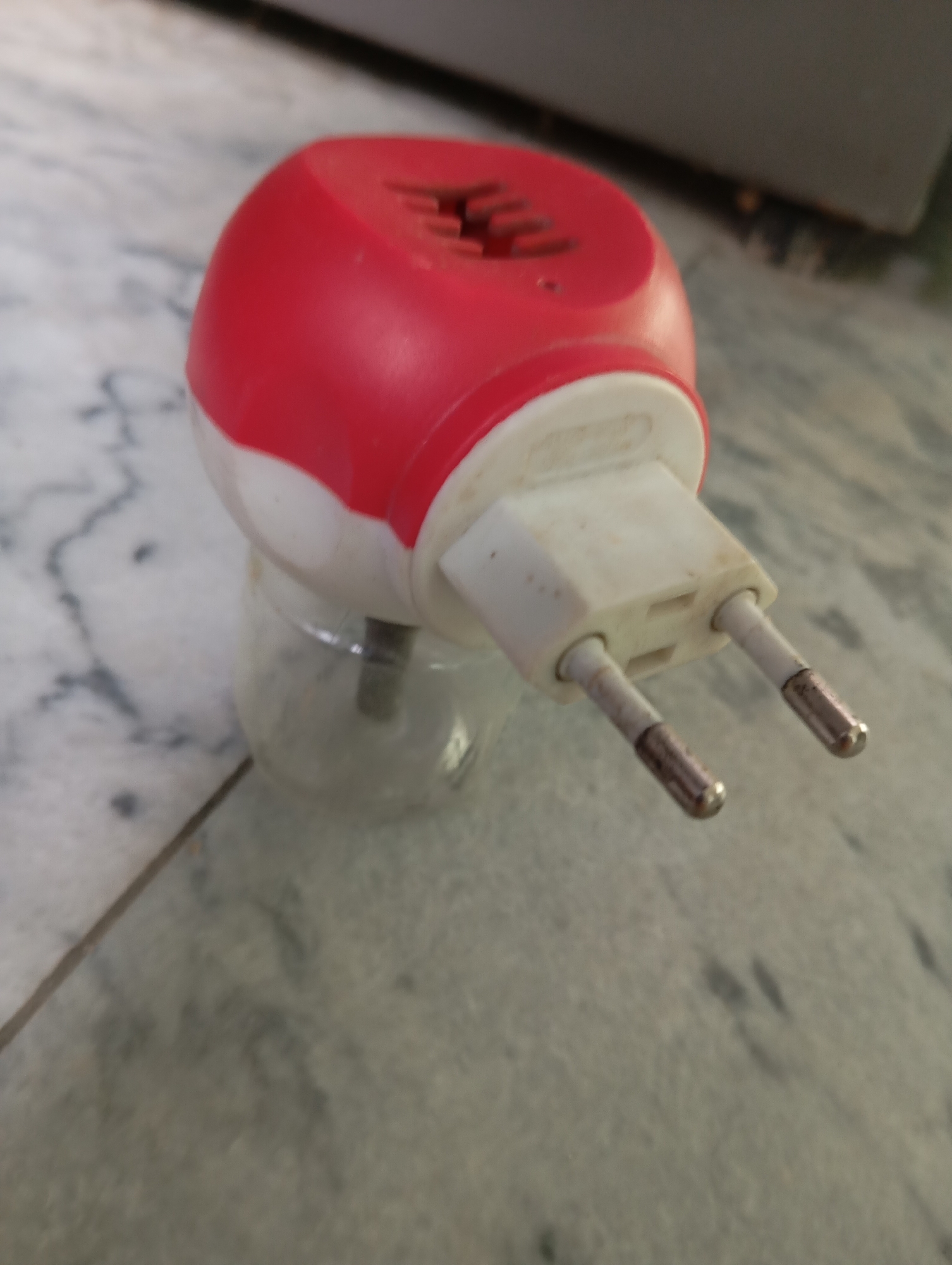 A red and white plug-in air freshener is shown on a marble surface. The air freshener has two metal prongs and a small glass bottle attached to the bottom, which contains a clear liquid. The surface appears to be a countertop or floor, with a faint pattern of grey veins running through the white marble. The lighting is soft and natural.