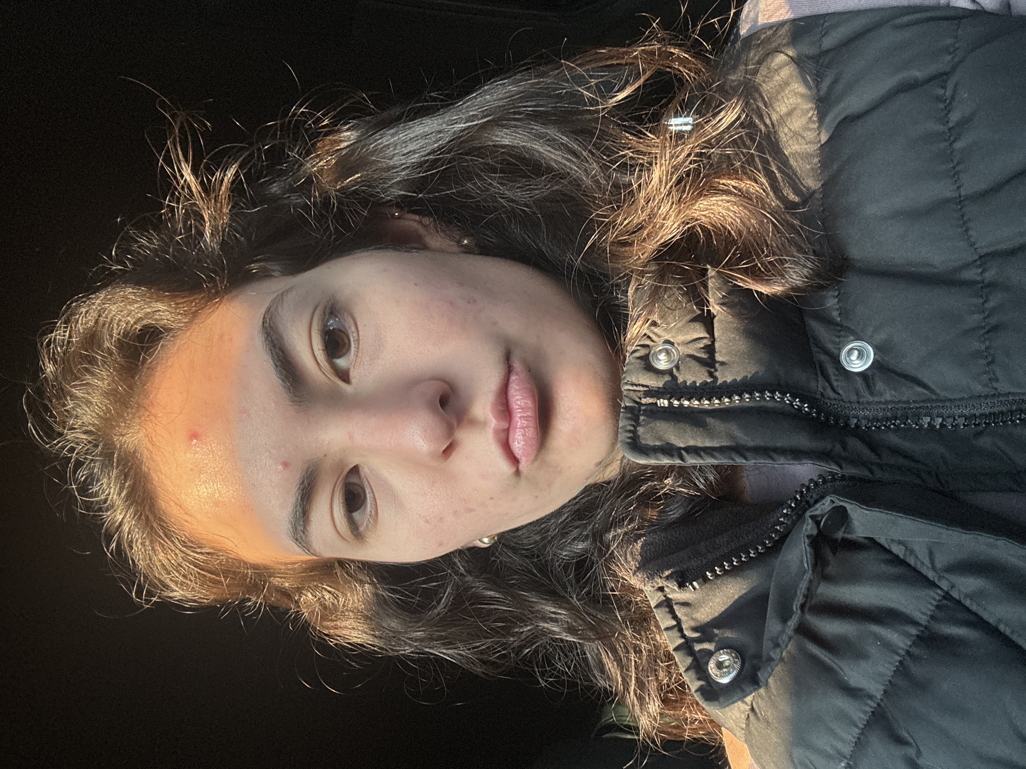 A person with curly brown hair is facing the camera, wearing a black padded vest over a light-colored top. The face is partially illuminated by sunlight, highlighting features such as the eyes, lips, and a blemish on the forehead. The background is dark and indistinct. There is no verifiable evidence to indicate this picture was taken in San Bernardino Tlaxcalancingo, Mexico.