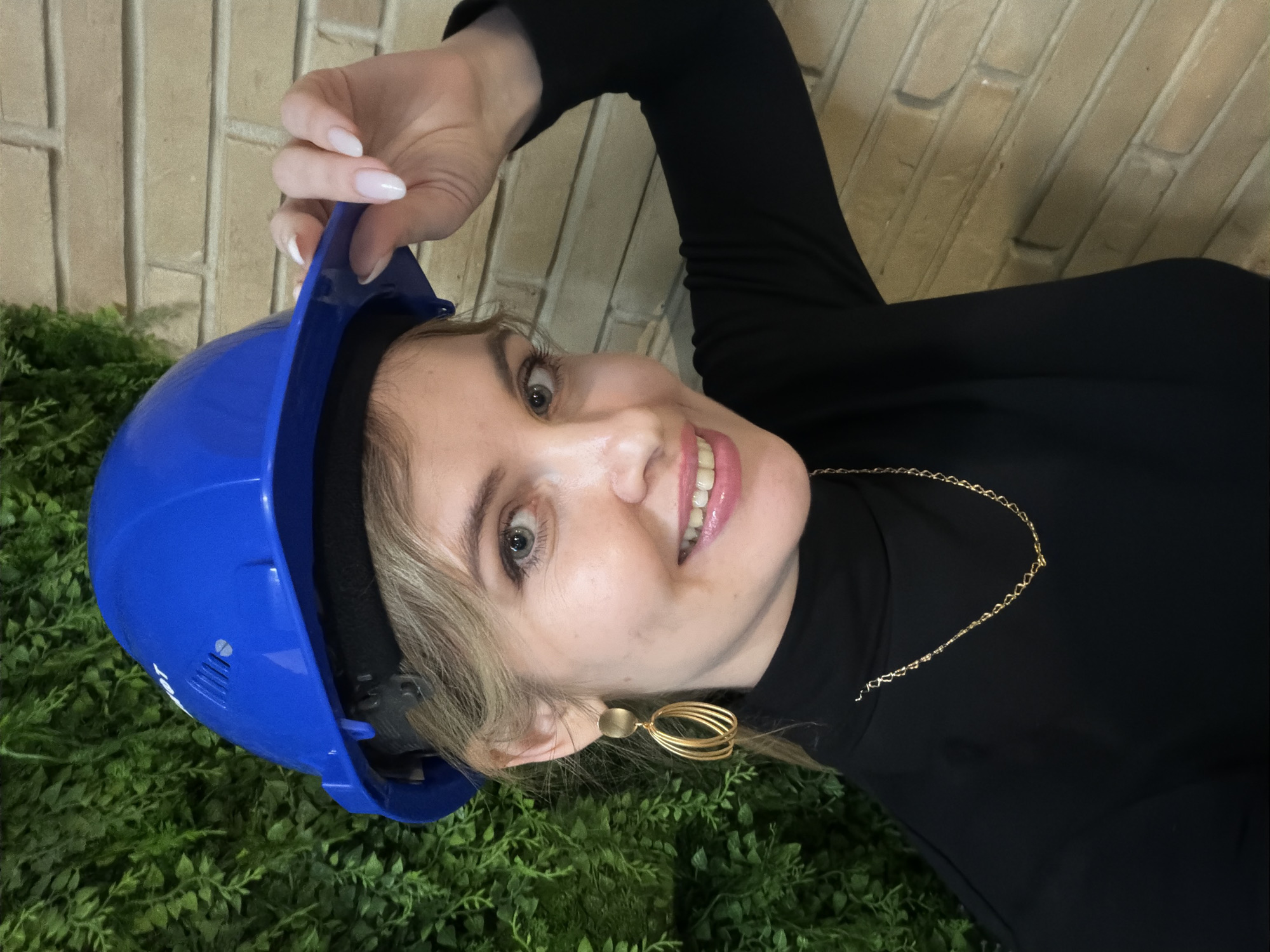 A woman is holding a blue hard hat while smiling at the camera. She is wearing a black top with a gold necklace and earrings. The background consists of a brick wall and some green foliage. The location information is not discernible.