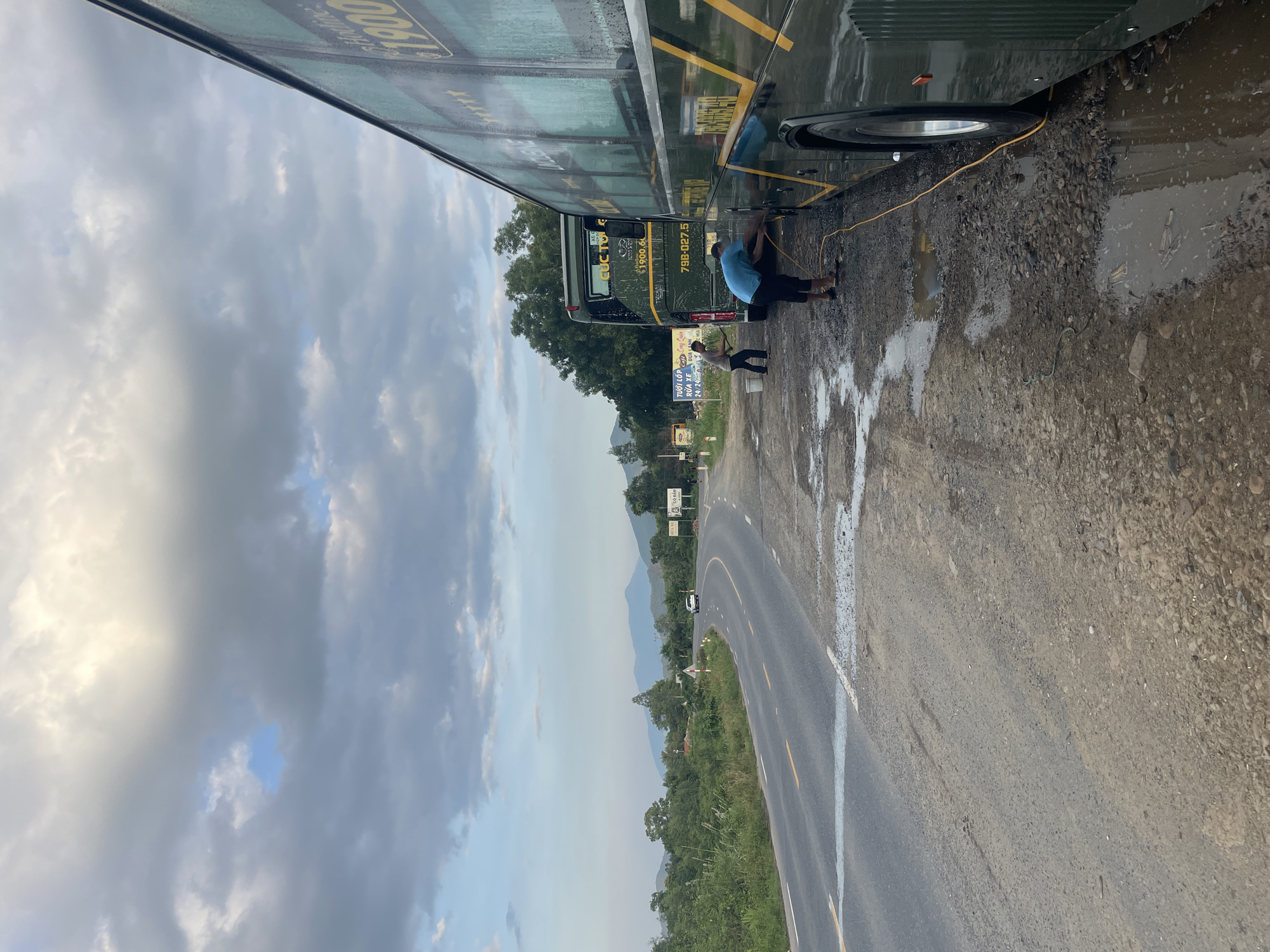 The image depicts a roadside scene in Tây Ninh Hòa, Vietnam, under a cloudy, late afternoon sky. Two large, dark green passenger buses are parked on a muddy and gravelly shoulder to the right of a two-lane asphalt road.
In the foreground, a person wearing a blue t-shirt and dark pants is bent over, actively cleaning the side of the closest bus with a yellow hose that extends across the muddy ground. Significant puddles of muddy water are visible around their feet and along the bus's side. This bus features yellow lettering and a "1900" hotline number on its dark green exterior.
Behind the first bus, a second identical dark green bus is parked, its rear section visible. A second person, wearing a grey t-shirt and dark pants, is standing near the rear of this bus, holding a cleaning tool, with buckets positioned nearby. The rear of this bus displays a license plate "79B-027.55" and the name "CUC TUNG". To the left of these buses, several commercial signs in Vietnamese are visible, one explicitly advertising "RỬA XE 24/24" (Car wash 24/24) and other services like cold coconut.
The asphalt road curves gently to the left and recedes into the distance, marked with a solid white line on its edge and a broken yellow centerline. A white car is observed driving away in the distance on this road. The immediate roadside on the left is lined with dense green vegetation, primarily trees and bushes. In the background, hazy blue mountains rise against the cloudy sky, which shows patches of lighter clouds and some glimpses of blue. The overall ground where the buses are parked is uneven, composed of dirt, gravel, and mud, indicating the ongoing washing activity and potentially recent wet conditions.