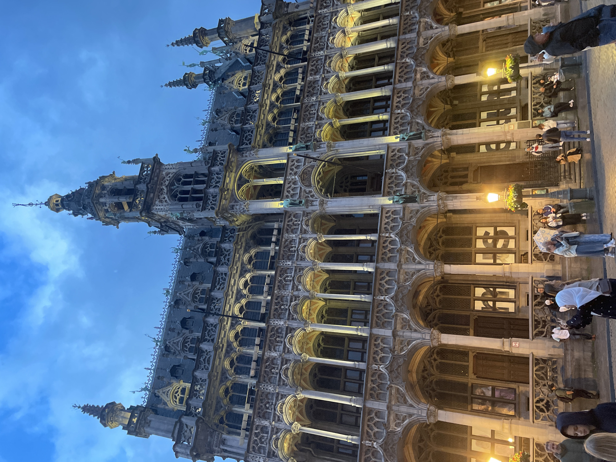 The image displays a majestic, ornate Gothic-style building, confirmed by context as the Brussels Town Hall, illuminated against a twilight sky. The grand structure features an abundance of architectural details, including numerous arched windows, intricate stone carvings, columns, spires, and turrets reaching towards the sky. Statues and what appear to be gargoyles are visible adorning the facade.
The scene is set at dusk or early evening, indicated by the deep blue sky with scattered clouds and the warm artificial lighting emanating from street lamps and floodlights that accentuate the building's elaborate features.
At the base of the building, several people are gathered. They appear to be tourists or locals, some looking up at the illuminated architecture, others engaged in conversation. One person in the foreground wears a dark hoodie, while another is seen holding an open umbrella. Decorative planters with flowers hang near the entrance, adding a touch of color. A small, red-bordered sign with a white 'H' is visible on a door at ground level, likely indicating an emergency exit or information point. The overall atmosphere is one of a bustling yet serene evening in a historic urban square.