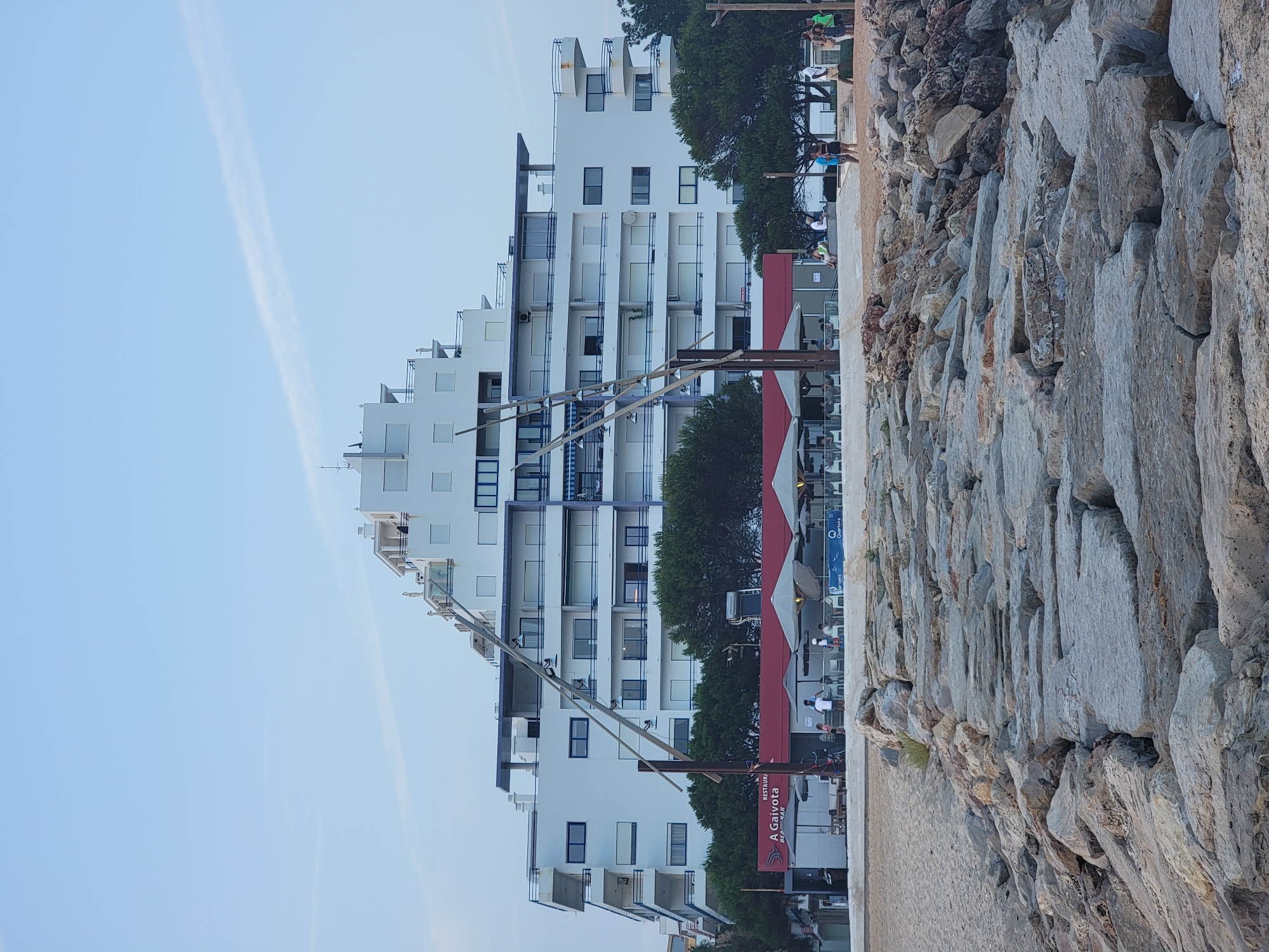 The image captures a view of a large, multi-story white building with a stepped pyramid-like structure at the top, overlooking a beach. In the foreground, a rocky breakwater extends into the water. A beach bar or restaurant with a red awning and outdoor seating is visible between the beach and the building. Several people are scattered around the beach bar area and on the sand. Trees line the area behind the beach bar. The sky is a pale blue with a faint cloud streak. The location is identified as Quarteira, Portugal.
