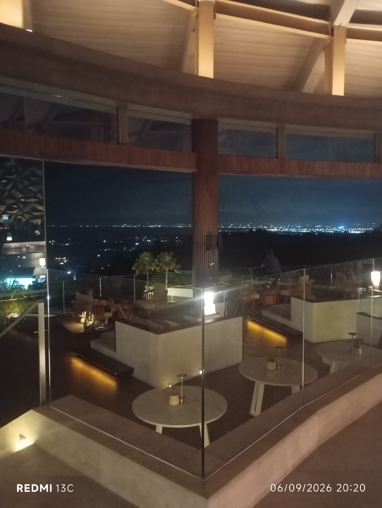 The image depicts an elevated outdoor seating area of a restaurant or lounge at night, overlooking a city landscape in Kuta Selatan, Indonesia. The architecture features a modern, open-air design with a curved wooden roof supported by visible beams. Below the roof, a large expanse of glass windows offers a panoramic view of the city lights spread across the horizon.
In the foreground, several glass partitions create separate seating sections. On a lower level, there are two round tables with chairs, illuminated by subtle lighting. One table has two small decorative items on it. In the middle ground, a raised platform contains lounge seating arrangements and small tables, with a potted plant as a central decorative element on one table. A person is partially visible seated on a lounge chair.
Further back, beyond the seating area and a protective railing, the city stretches out, dotted with numerous lights. The night sky is dark, with some faint clouds visible. The overall ambiance is that of a sophisticated establishment designed for enjoying the nighttime view. The bottom of the image includes a timestamp and a device model, "06/09/2026 20:20 REDMI 13C".