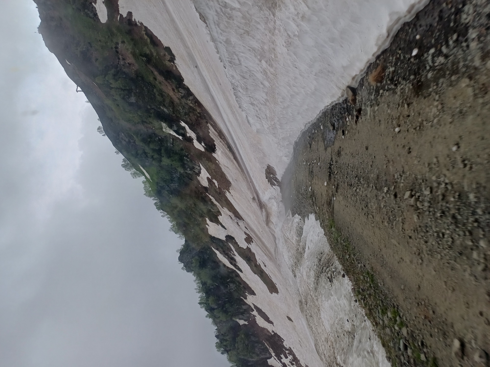 The image shows a mountain road in Estosadok, Russia, with patches of snow on the slopes. The road is unpaved and covered with gravel. The sky is overcast, and there is some mist or fog in the distance. The mountain slopes are covered with sparse vegetation, with trees and bushes visible higher up the mountain. There are no people or animals visible in the image. The overall impression is of a rugged and natural landscape, possibly in the early stages of spring or late autumn.