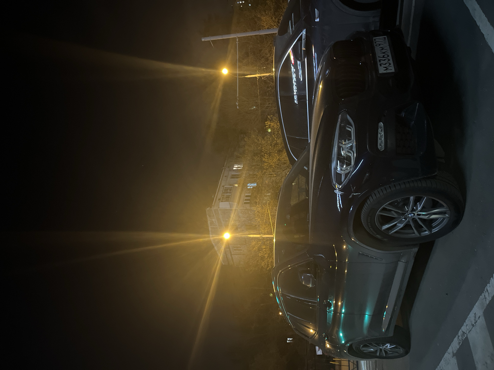 A nighttime image shows two dark-colored BMW SUVs parked next to each other. The car in the foreground is angled towards the viewer and occupies most of the right side of the frame. Its headlights are visible, and its license plate reads "M336KM977". The second SUV is partially visible behind the first one. The scene is illuminated by streetlights, with one directly behind the cars and others visible further down the street. In the background is a building with illuminated windows, suggesting it is residential. The cars are parked on a paved surface marked with white lines. Based on the license plate, the location is likely Moscow, Russia.