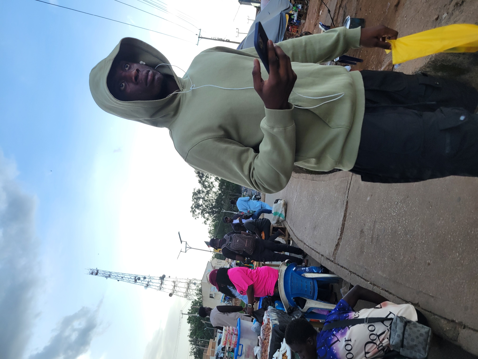 A young Black man wearing a light green hoodie and black cargo pants walks on a paved sidewalk, holding a dark-colored smartphone in his right hand and a bright yellow plastic bag in his left. He is looking to his right, and white earbuds are visible in his ears. The sidewalk runs alongside a busy market, with various stalls and people visible in the background. A tall telecommunications tower is prominent in the distance. The scene appears to be in Jos, Nigeria, given the presence of a typical market environment and infrastructure common to the region.