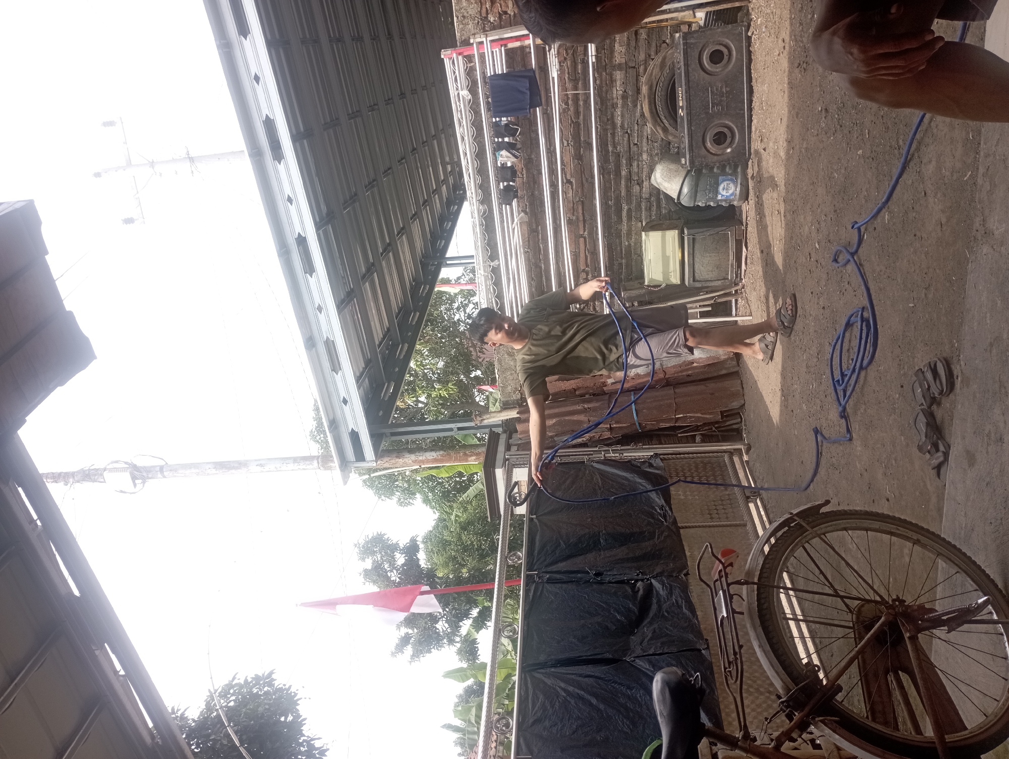 The image depicts an outdoor scene in Maos Kidul, Indonesia. A young man wearing a green t-shirt and gray shorts stands in the center, holding a blue hose. He appears to be in the process of coiling or manipulating the hose. To his left, the rear wheel of a rusty bicycle is visible. In the background, a clothesline with several items of clothing hangs on a brick wall. Various objects, including what appear to be old electronics and tires, are scattered on the ground. A red and white Indonesian flag is prominently displayed on a pole to the left of the man. The overall environment suggests a residential or utility area with a corrugated metal roof overhead.