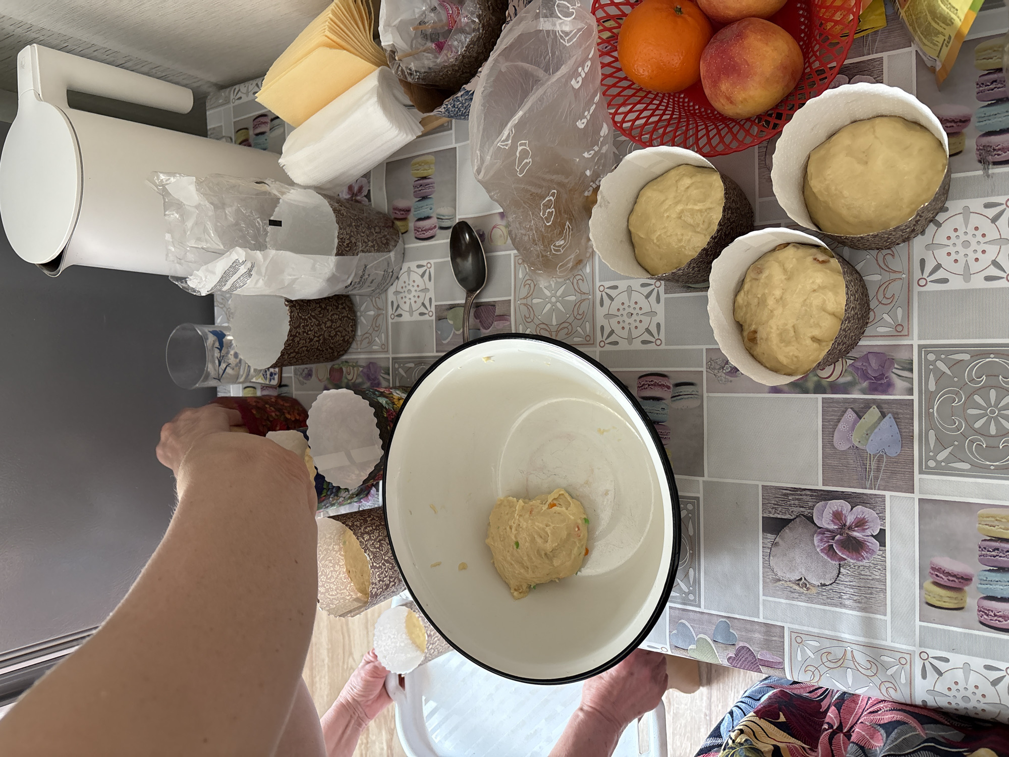 A photograph taken in Ust-Ilimsk, Russia, shows a woman's arms and hands preparing dough, likely for baking Easter cakes. The woman is holding a paper baking cup, presumably to fill it with dough from a white mixing bowl. Three other baking cups already contain dough, and there's a red plastic basket holding fruit (oranges and peaches). Other items are visible on the countertop, including an electric kettle, a spoon, a bag of ingredients, and packaged goods. The countertop has a decorative pattern featuring flowers and macarons.