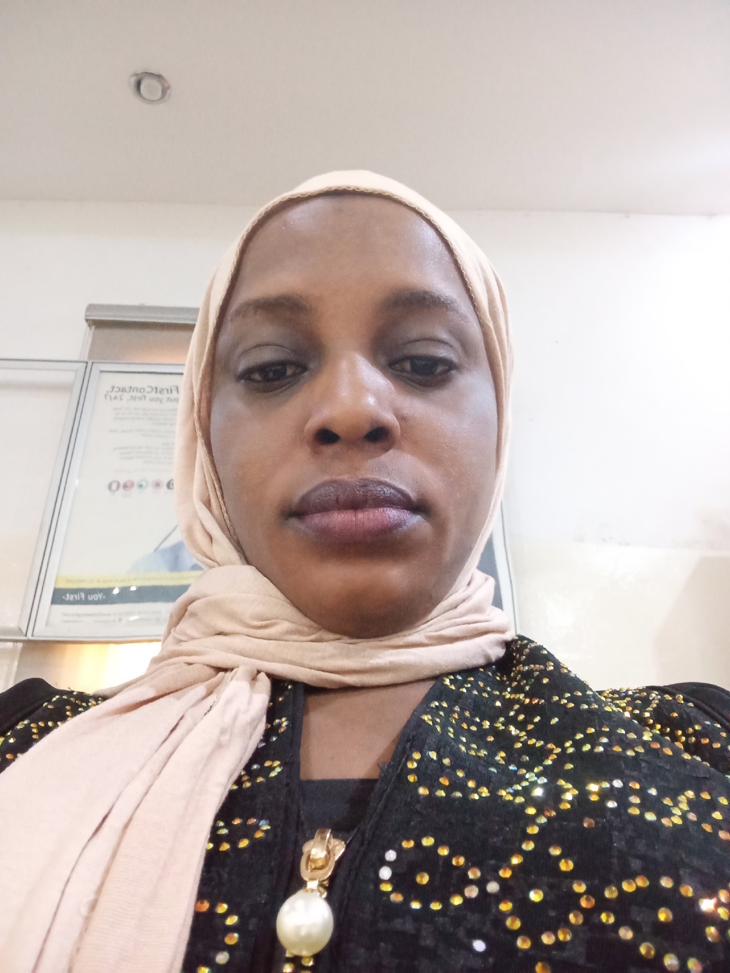A close-up selfie captures an adult woman from the chest up, centered in the frame, in what appears to be an indoor setting in Kaduna South, Nigeria. She has a dark skin tone and a neutral, composed expression, looking directly at the camera. She wears a light beige or nude-colored hijab draped over her head and neck. Her attire includes a black garment, possibly a shirt or jacket, heavily embellished with numerous small, sparkling gold or yellow accents, resembling sequins or beads, scattered across the fabric. A distinct gold-colored pendant, featuring a prominent, large white pearl, hangs from her neck.
The background consists of light-colored walls and a ceiling. To the woman's left, a framed sign or poster is partially visible, appearing mirrored due to the selfie perspective. Identifiable text on this sign includes "First Contact" and "You First," along with a string of numbers that starts with "080". On the ceiling in the upper left quadrant, a circular, recessed light fixture is visible. The lighting is bright and even, suggesting an indoor environment during the day. The scene depicts a straightforward portrait, with no specific activity other than posing for the photograph.