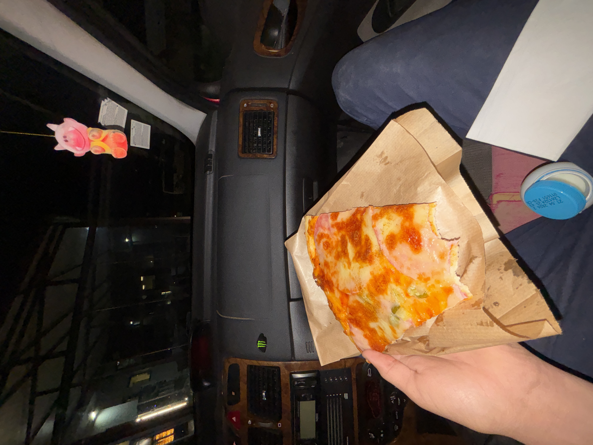 Here is a forensic description of the image:
A slice of pizza sits in a brown paper bag, held by a fair-skinned hand. The hand is positioned inside a vehicle, with a dark gray dashboard directly behind the pizza. Part of a seat covered in navy blue fabric is visible to the side. To the left is the vehicle's window, showcasing a dark night scene outside. Lights from buildings are scattered across the horizon. A pink pig-shaped decoration hangs from the top of the window. A blue-lidded container sits on the seat.