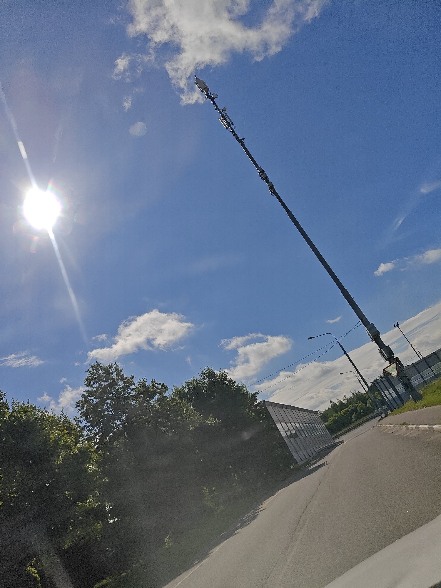 In Pushkino, Russia, a cell tower stands tall against a bright blue sky dotted with white clouds. The sun shines intensely to the left of the frame, casting a lens flare. The tower is positioned diagonally, extending from the bottom right of the image towards the top right. Atop the tower, several antennas are visible. Below the tower, a road curves to the right, bordered by a sound barrier made of metal and glass. Lush green trees line the left side of the road, and a traffic light is visible in the distance.